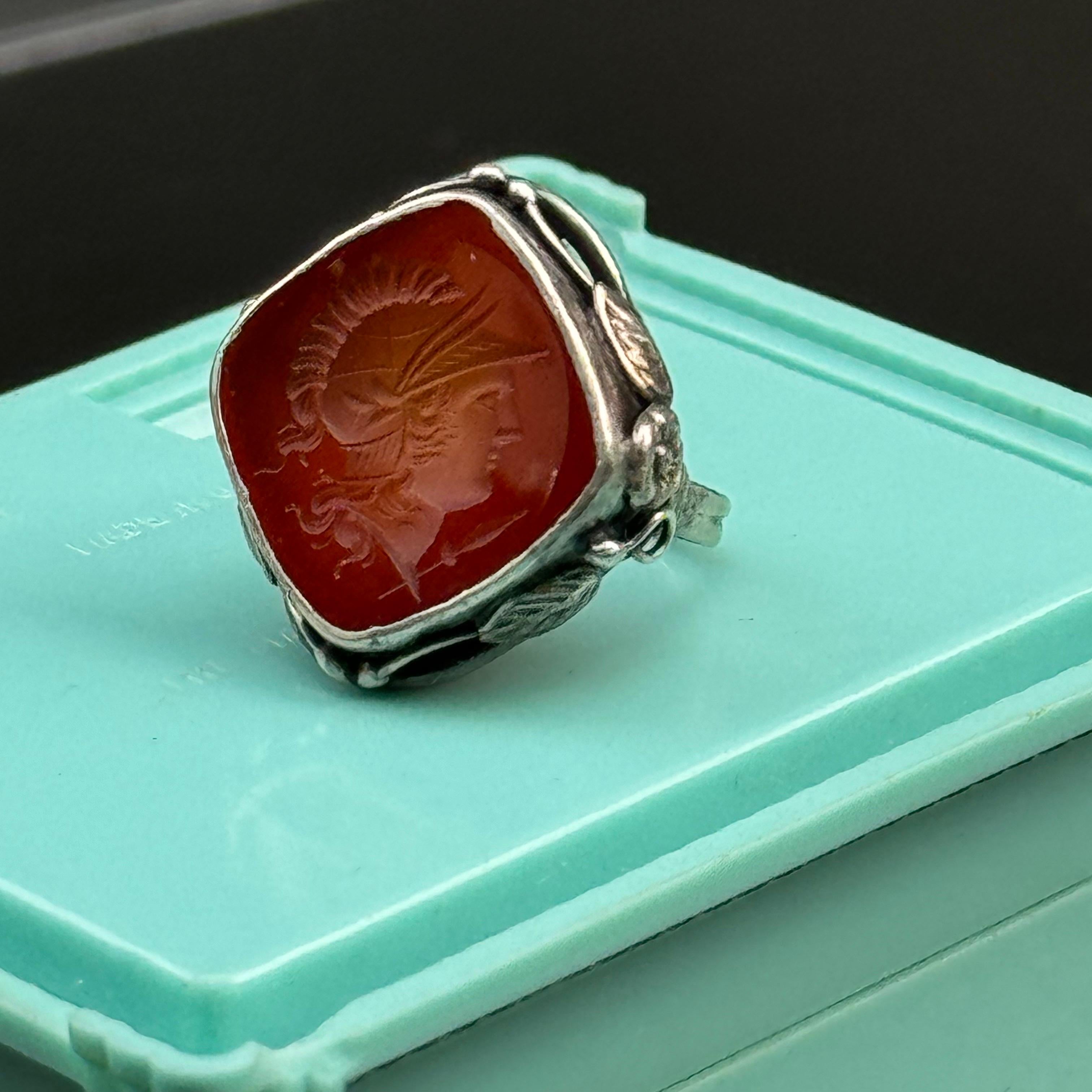 Adéntrate en la elegancia de antaño con este exquisito anillo vintage, que tiende un hermoso puente entre los estilos Arts & Crafts y Art Nouveau. Este llamativo anillo presenta una cornalina central tallada y engastada en un marco de diseño floral