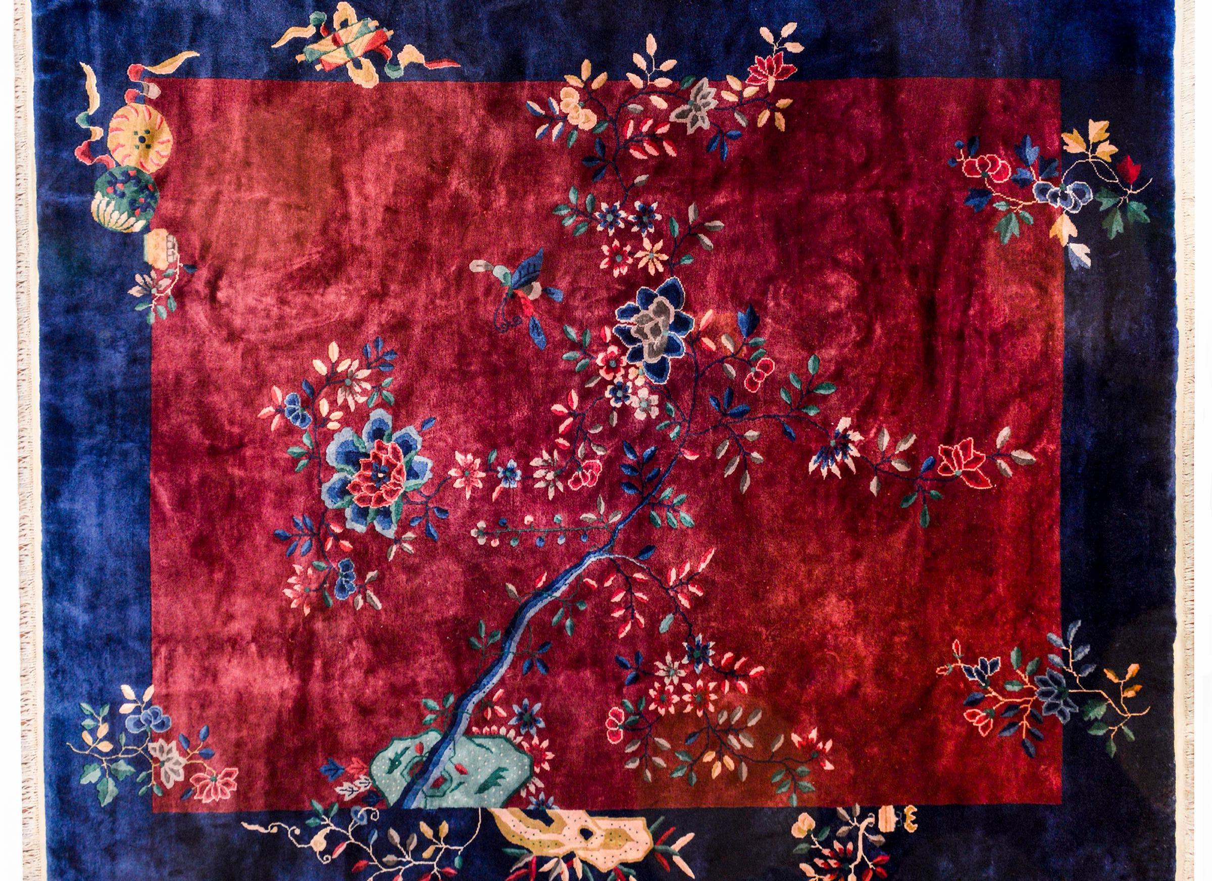 A gorgeous Chinese Art Deco rug with a deep shimmering cranberry field with a wide dark indigo border. A scholar's rock appears at the bottom, with a single branch with multiple species of flowers including peonies, chrysanthemums, and prunus.
