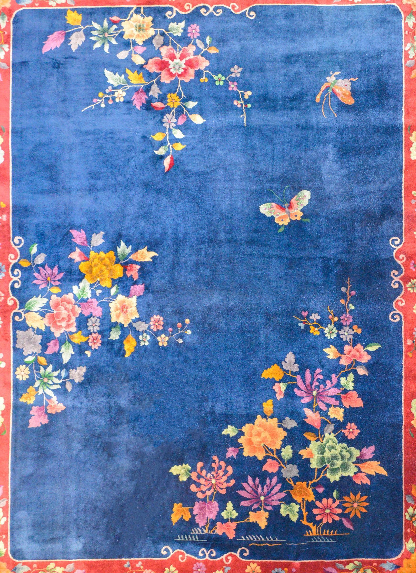 A gorgeous early 20th century Chinese Art Deco rug with a bold dark indigo field surrounded by a wide cranberry border with scrolled lines. Clusters of bight multicolored peonies, chrysanthemum, and cherry blossoms dot the field, while a pair of