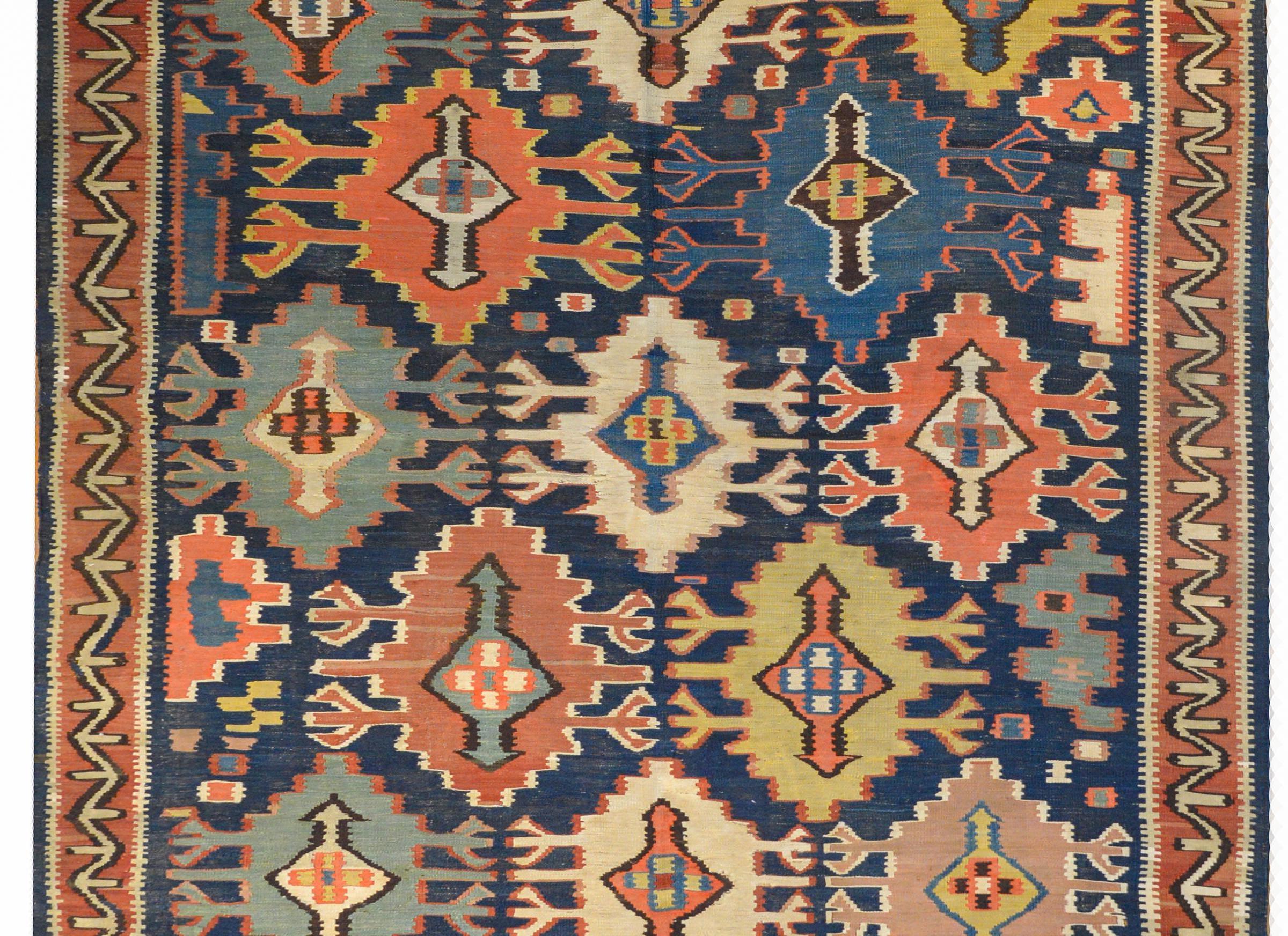 A gorgeous early 20th century Kuba Kilim rug with large boldly patterned stylized floral shapes rendered in myriad colors on a brilliant indigo background. The border is simple with a geometric pattern woven in crimson, black, and white vegetable