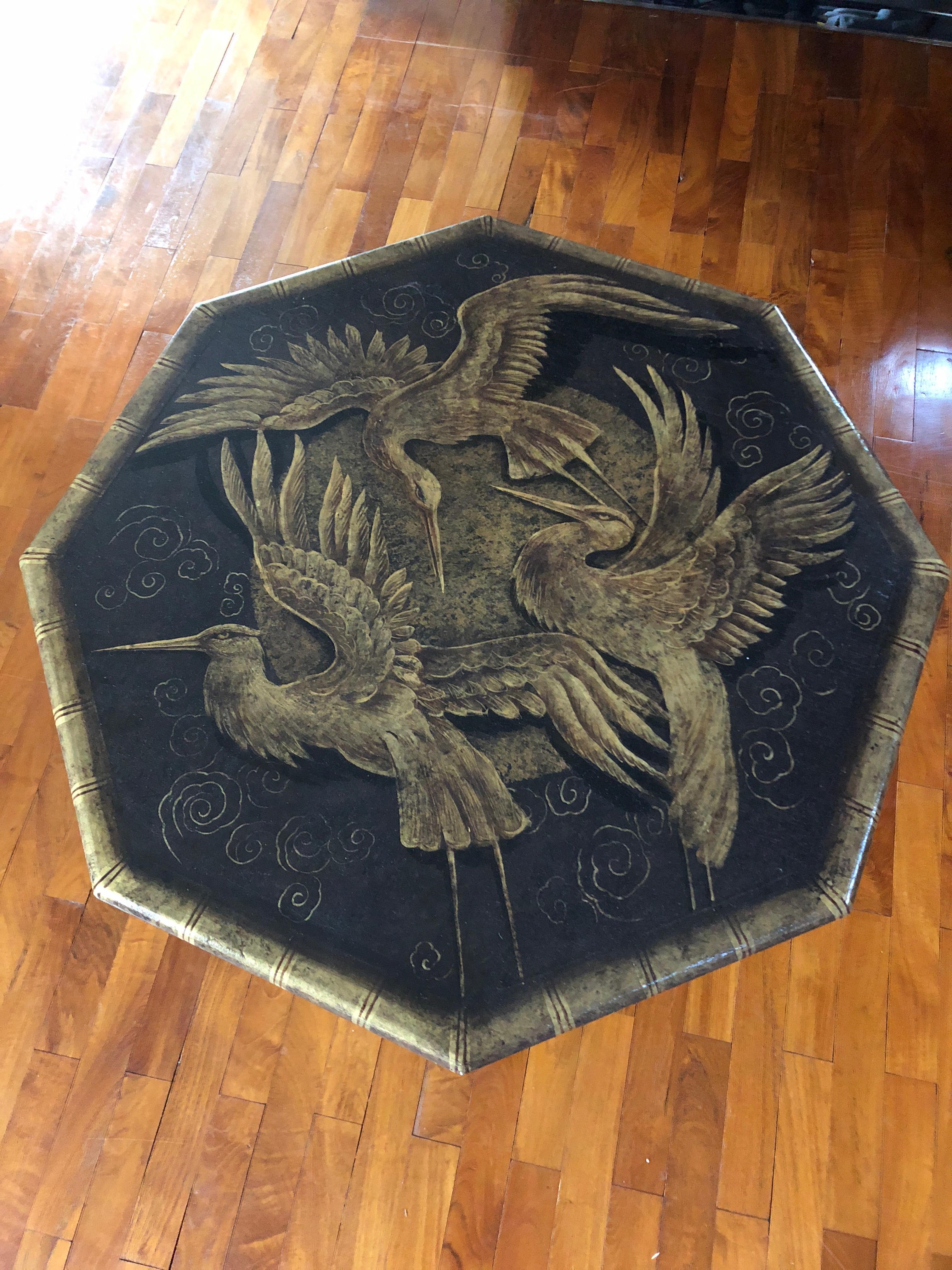 A very richly made octagonal side table having a painted top featuring cranes or birds and foliage in gold against black. The edges look like painted bamboo, and the iron tripod base is thin and elegant, but heavy and solid.