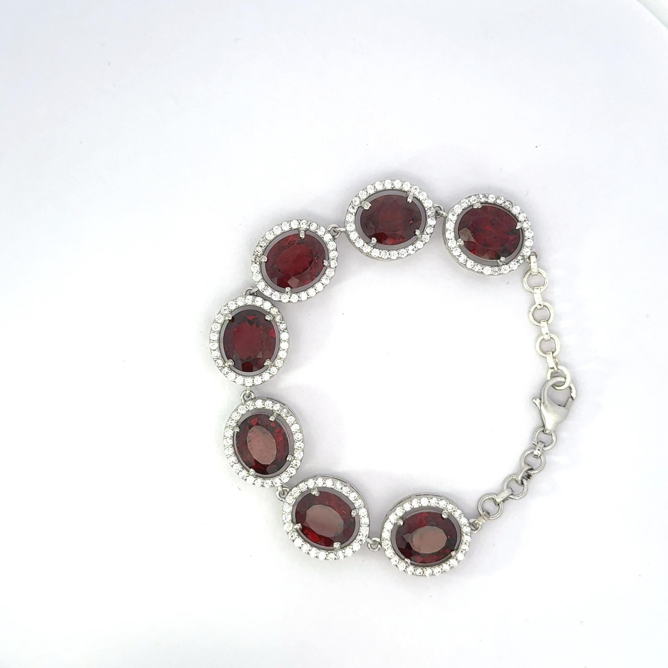 Esta llamativa pulsera de eslabones con halo de circonitas blancas y rojas profundas de corte ovalado es de plata de ley 925. Con siete piedras de color granate de talla ovalada ricamente saturadas, cada una rodeada por un halo de brillantes gemas