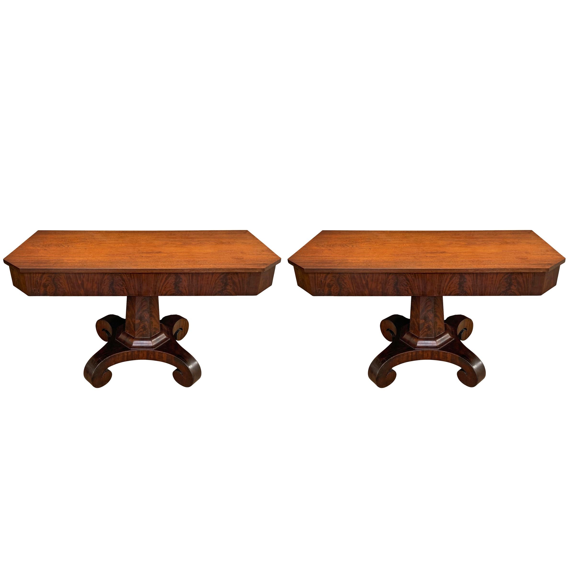 A gorgeous pair of early 19th century American Empire crotch mahogany veneer console tables with classical architectural details including tapered octagonal columns and hefty scrolled feet with their original casters, with tops made from solid