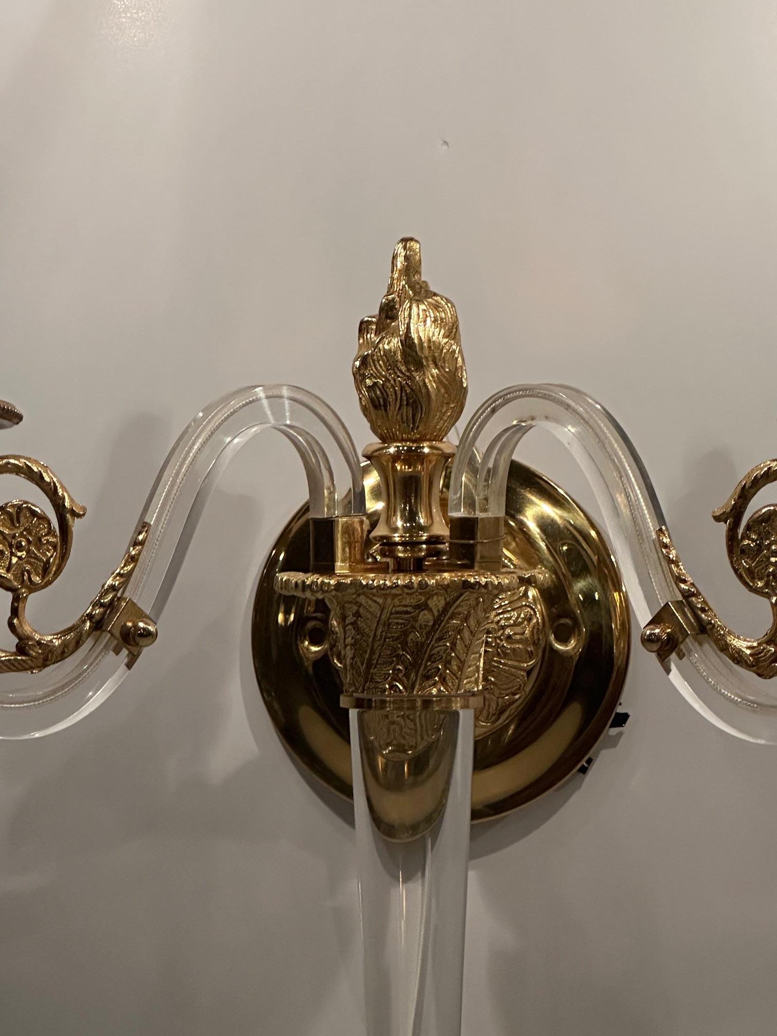 Glamorous Hollywood Regency style lucite and brass wall sconces having neoclassical elements including central flame finial and rosettes on the arms, two arms each