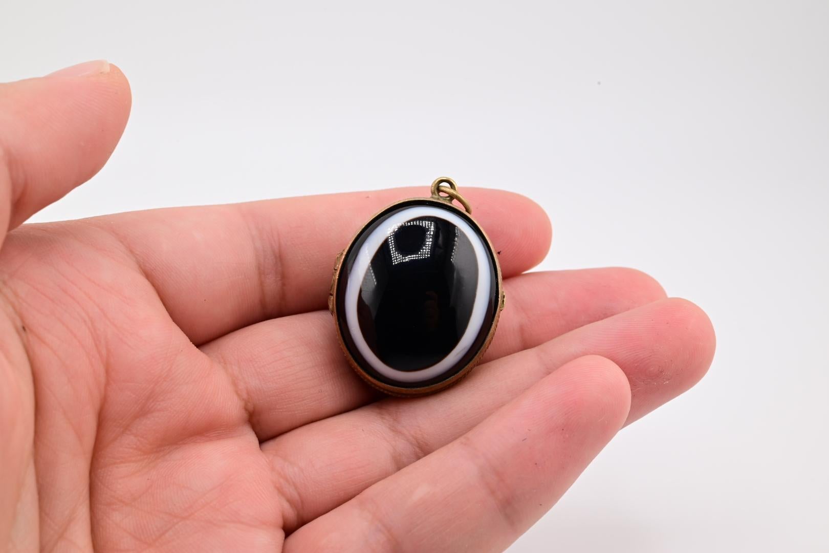 This is a striking pendant featuring a two black stone agate with a smooth, glossy surface. The stone is encircled by a subtle white band, creating a bold contrast. The locket pendant is set in a simple, elegant metal frame with a small loop for