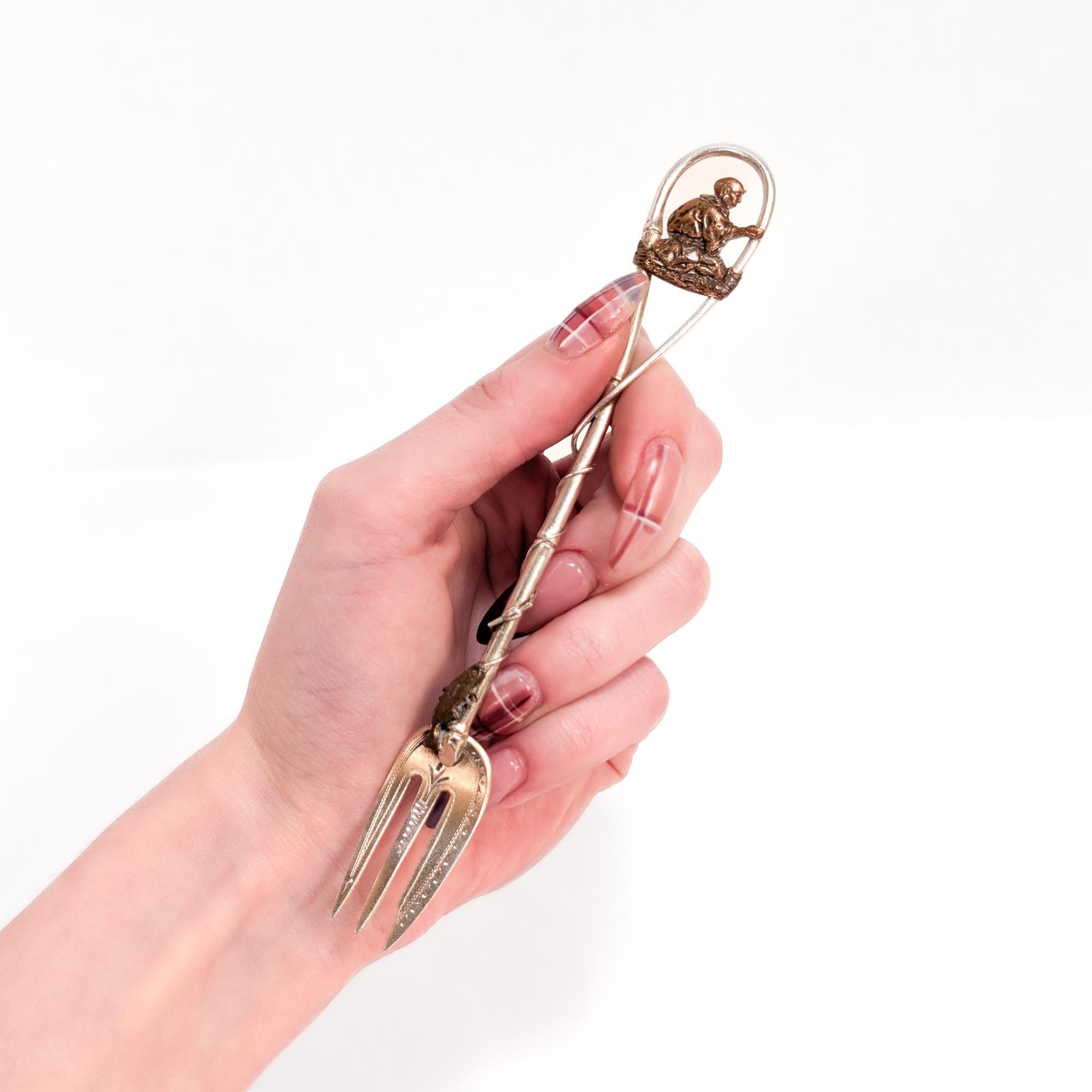 Fourchette à cocktail figurative de la période esthétique.

Par Gorham. 

En argent sterling et autres métaux avec des dents dorées.

Dans le style japonais, avec un pêcheur masculin en cuivre assis sur un tronc en haut de la poignée et un poisson