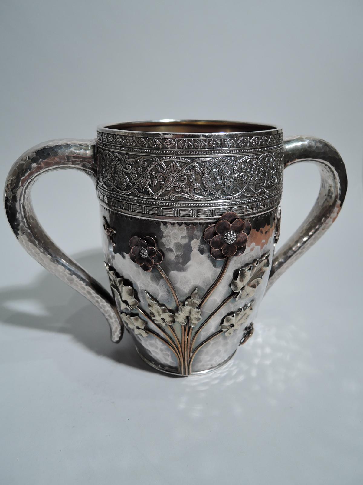 Japonesque sterling silver and mixed metal cup. Made by Gorham in Providence in 1882. Curved sides and s-scroll side handles. Allover honeycomb hand hammering. At top is wide band with stylized ornament: rinceaux between beaded, rosette, and