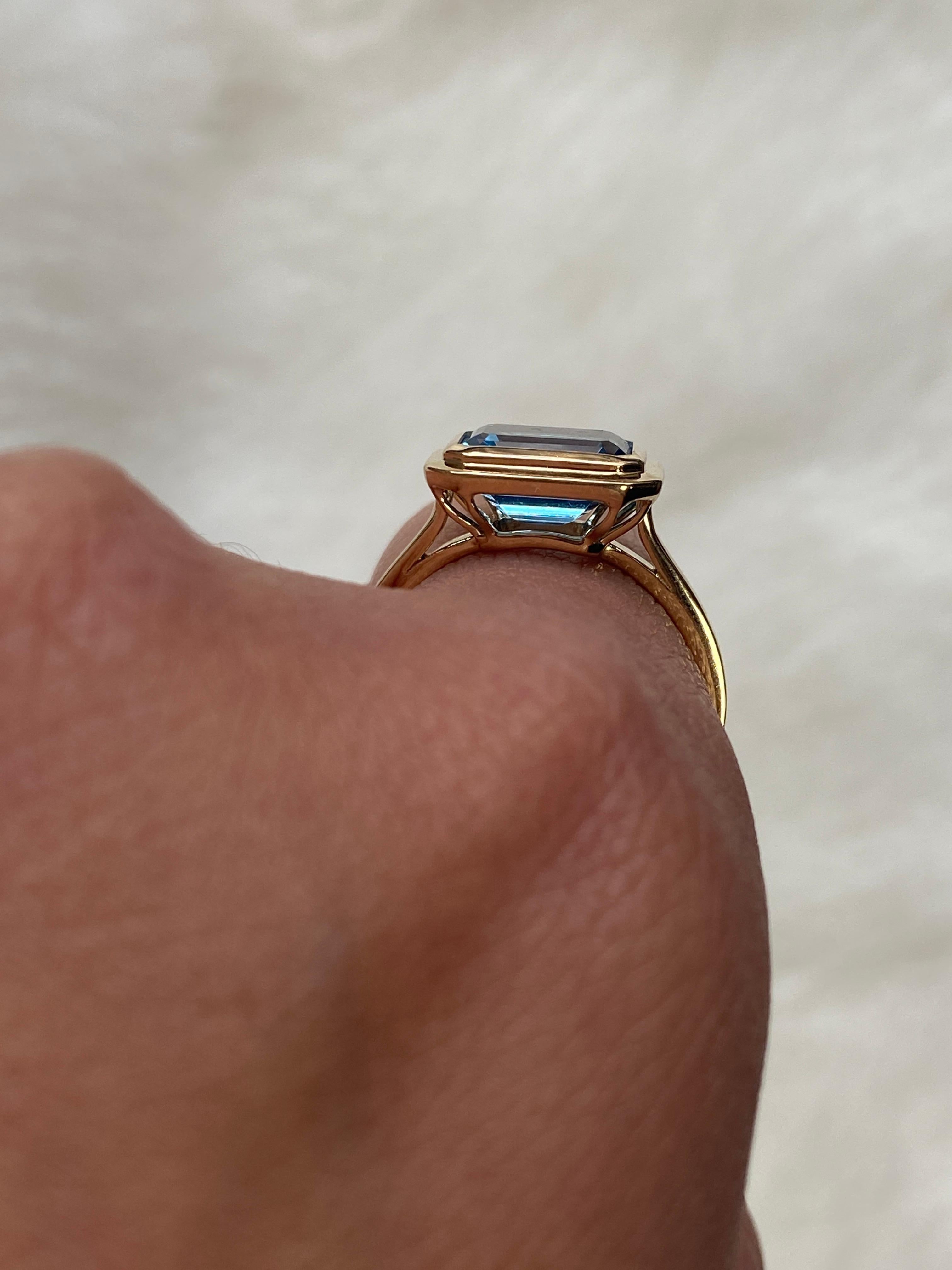 This Blue Topaz Emerald Cut Bezel Set Ring in 18K Yellow Gold is a sleek piece from the 'Manhattan' Collection. It features a stunning emerald-cut Blue Topaz stone set in a 18K yellow gold bezel. This ring embodies modern elegance, offering a touch