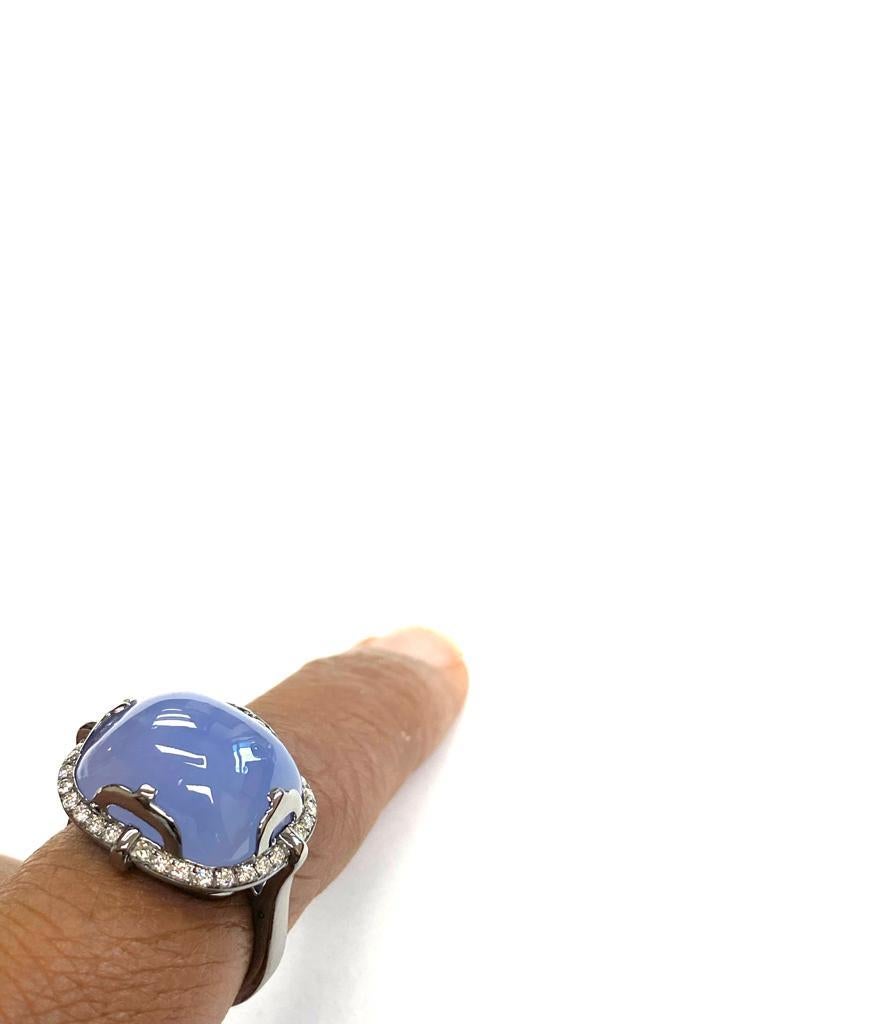 This Blue Chalcedony East-West Cushion Cabochon Ring with Diamonds in 18k White Gold and Rhodium is a stunning piece of jewelry from the 'Rock N Roll' collection. The ring features a vibrant blue chalcedony stone cut into a cushion shape and set