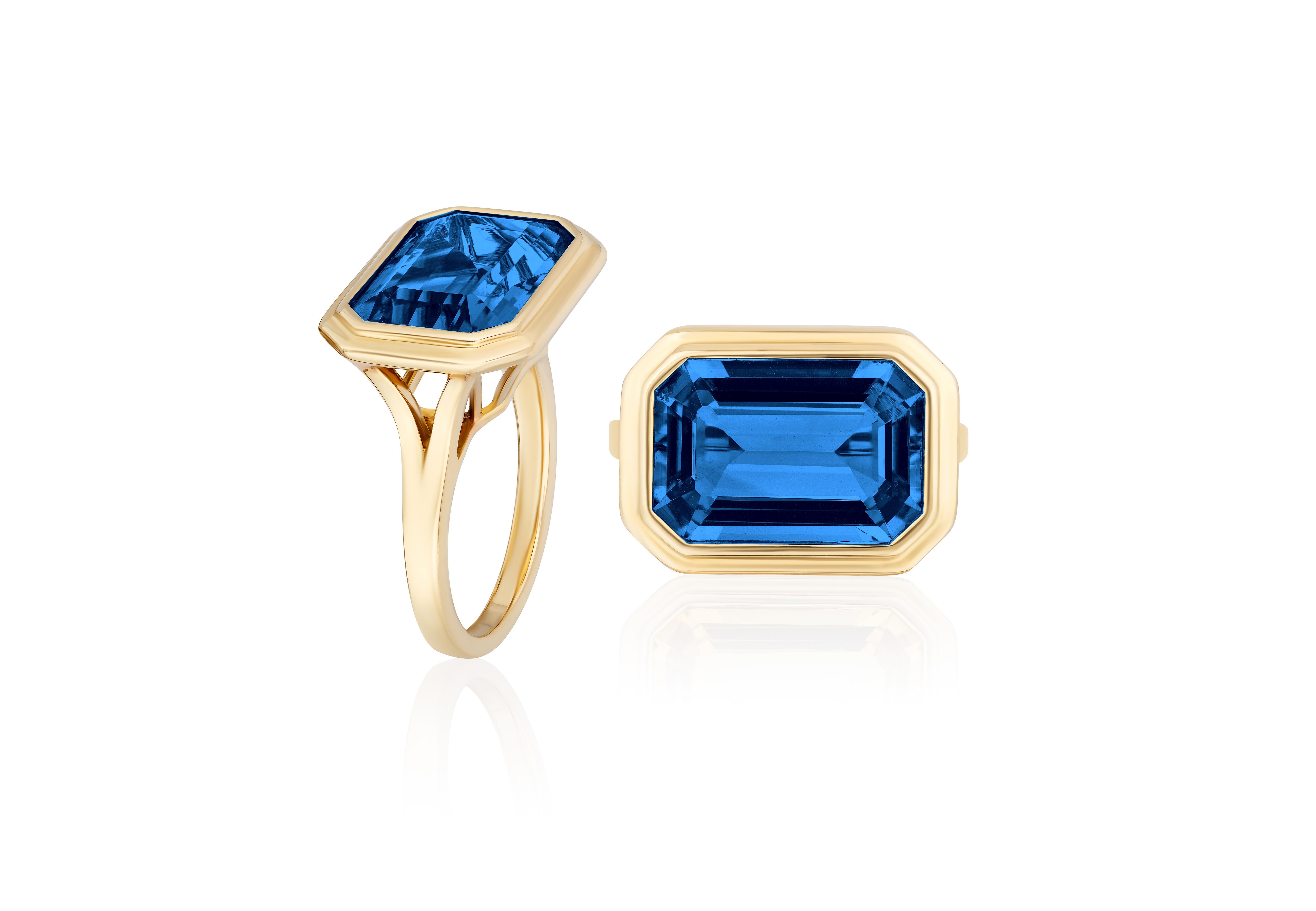 The East-West London Blue Topaz Emerald Cut Bezel Set Ring is an elegant piece of jewelry from the 'Manhattan' Collection. The ring is crafted from 18K yellow gold and features a beautiful London blue topaz in an emerald cut, set horizontally in a