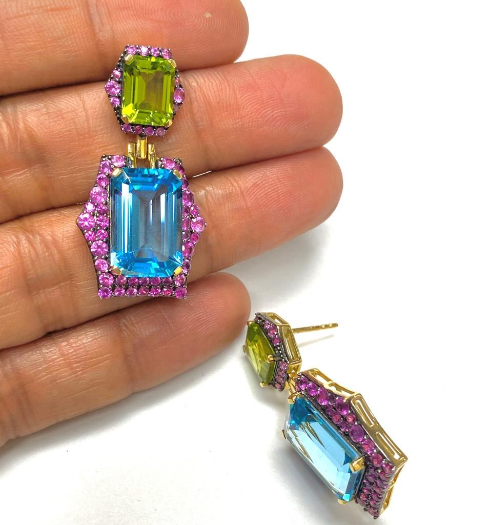 These Blue Topaz & Peridot Emerald Cut Earrings with Pink Sapphire in 18K Yellow Gold And Black Rhodium are a stunning piece of jewelry from the 'Rainforest' Collection. The earrings feature beautiful emerald cut blue topaz and peridot gemstones,