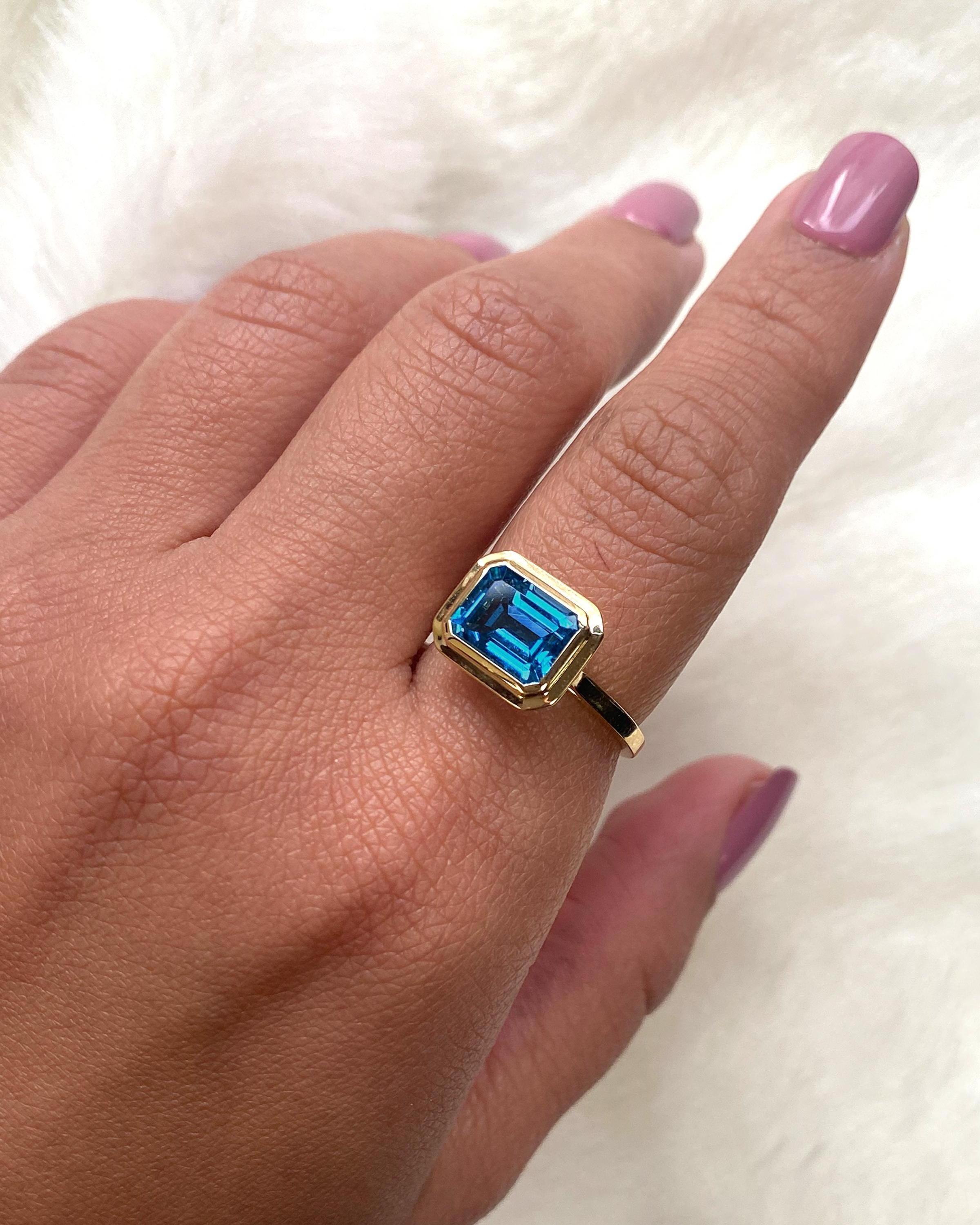 This London Blue Topaz Emerald Cut Bezel Set Ring in 18K Yellow Gold is a sleek piece from the 'Manhattan' Collection. It features a stunning emerald-cut London Blue Topaz stone set in a 18K yellow gold bezel. This ring embodies modern elegance,