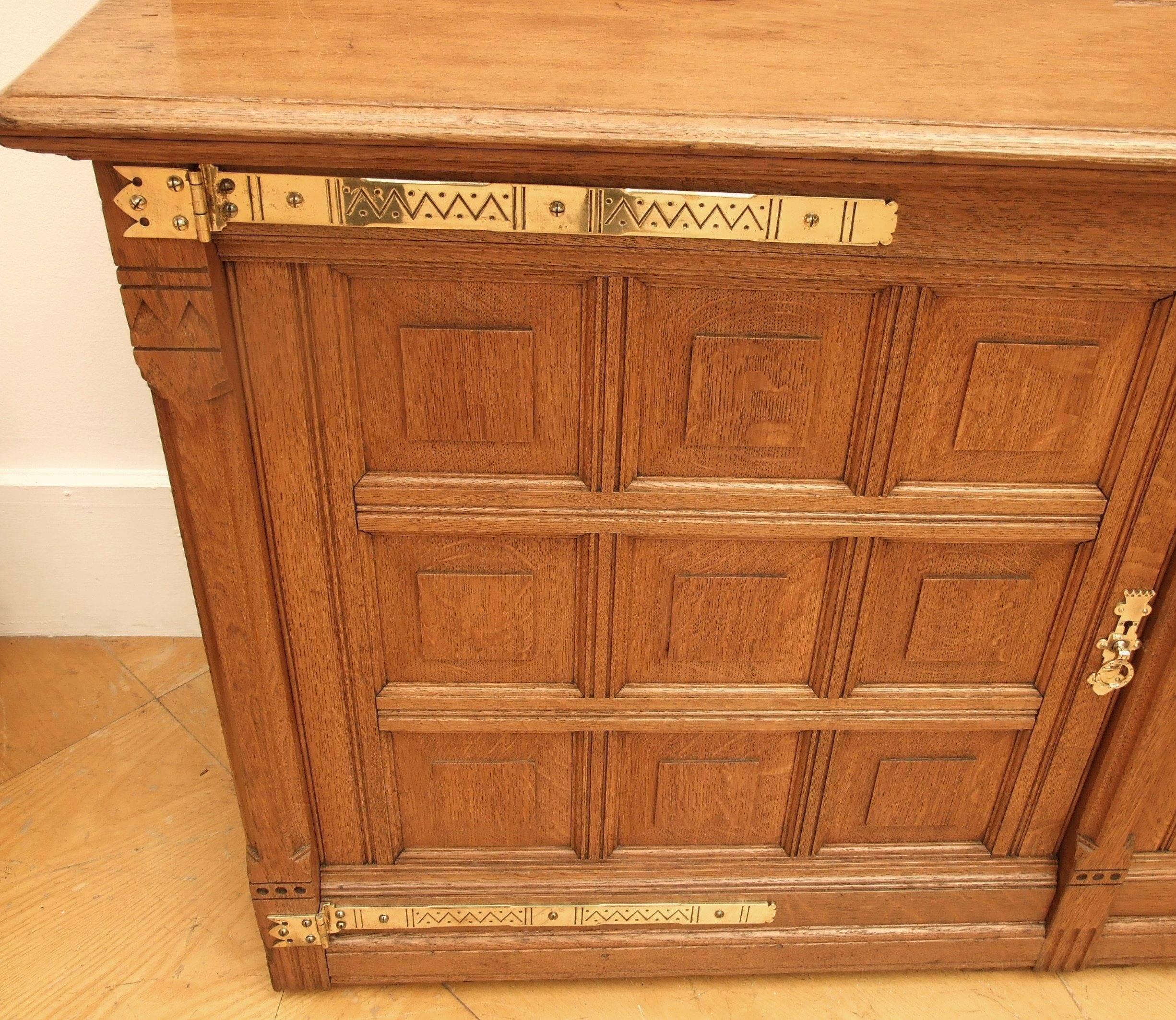 Attributed to Richard Norman Shaw An Architectural, Muscular Gothic Revival, oak bookcase, two glazed doors over a two door cupboard, carved, stylised foliage to the cornice, chevron carving and stepped chamfering to the frame, coved cornice with