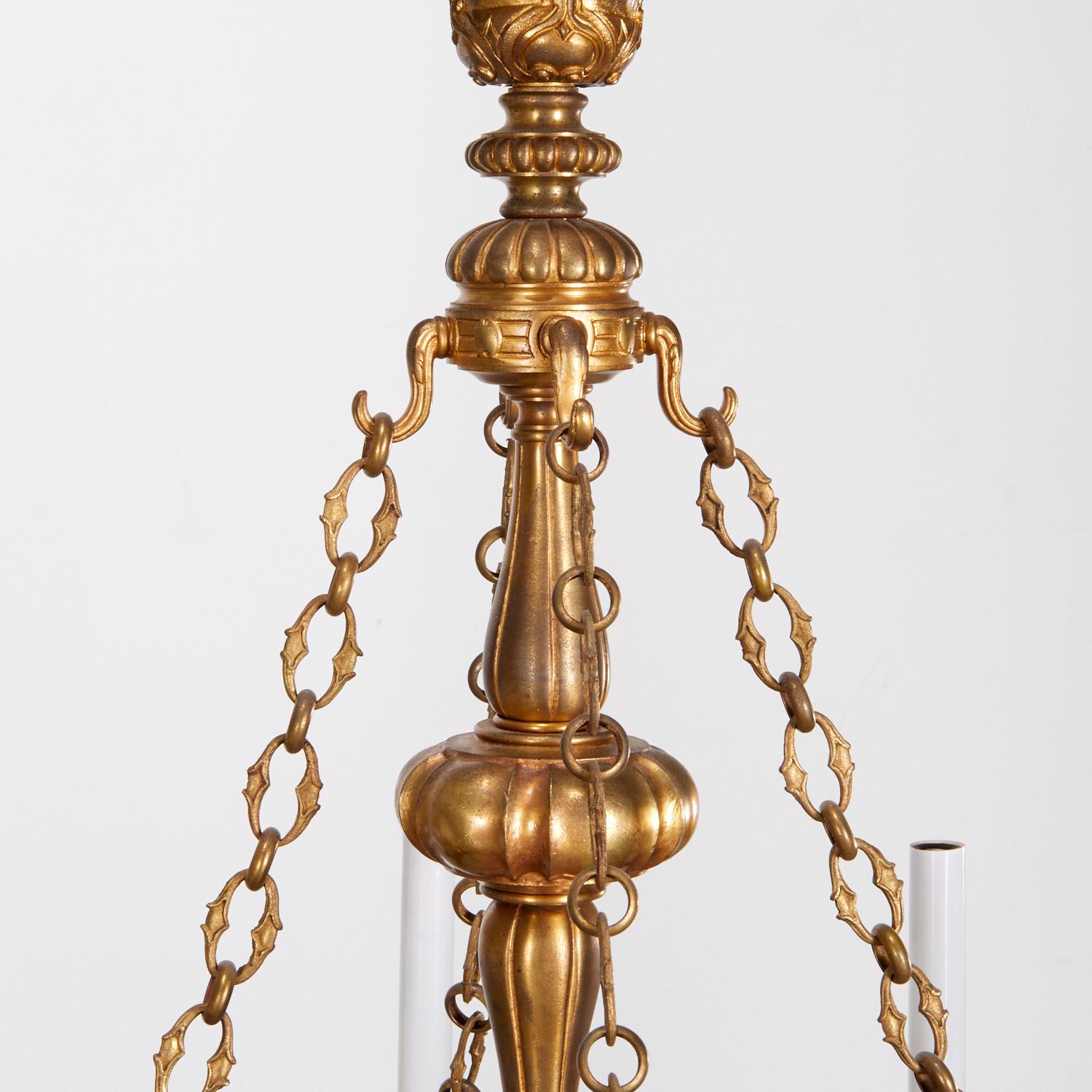 Lampadario in ottone in stile antico del 20° secolo con una ricca finitura dorata invecchiata. La patina aggiunge profondità e carattere, conferendo un aspetto autentico e vintage. Il lampadario ha una struttura circolare con intagli dettagliati e