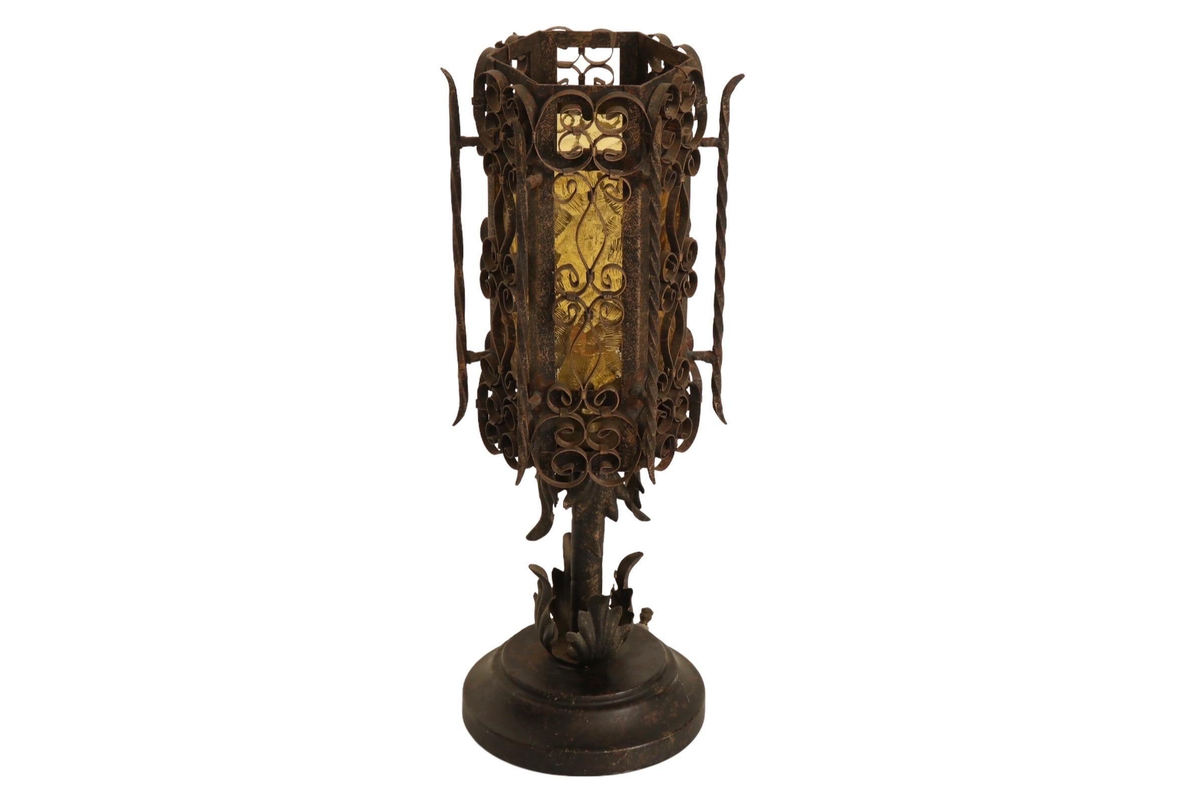 A Gothic lantern style hexagonal table lamp. Textured amber glass panes on each side sit behind scrolled elements and twisted wrought iron posts decorate the corners of the frame. The lamp is supported by a single iron column with four acanthus