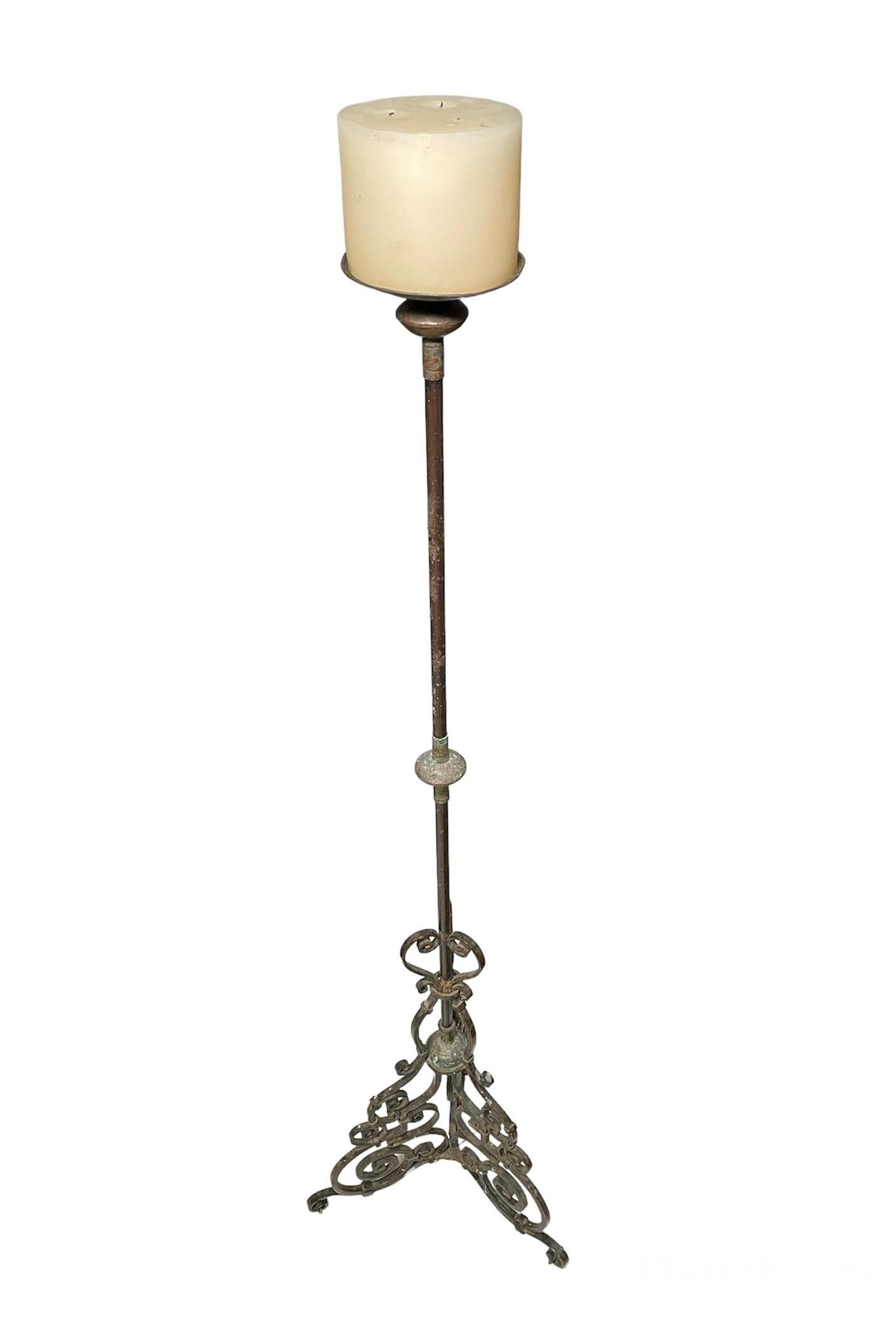 This is a tall wrought iron candle holder/torchere that ends in a tripod base. Each leg of this base is adorned with large scrolls that start in the center stand forming a heart and finish in each leg. One wide round candle sits over the candle pan.