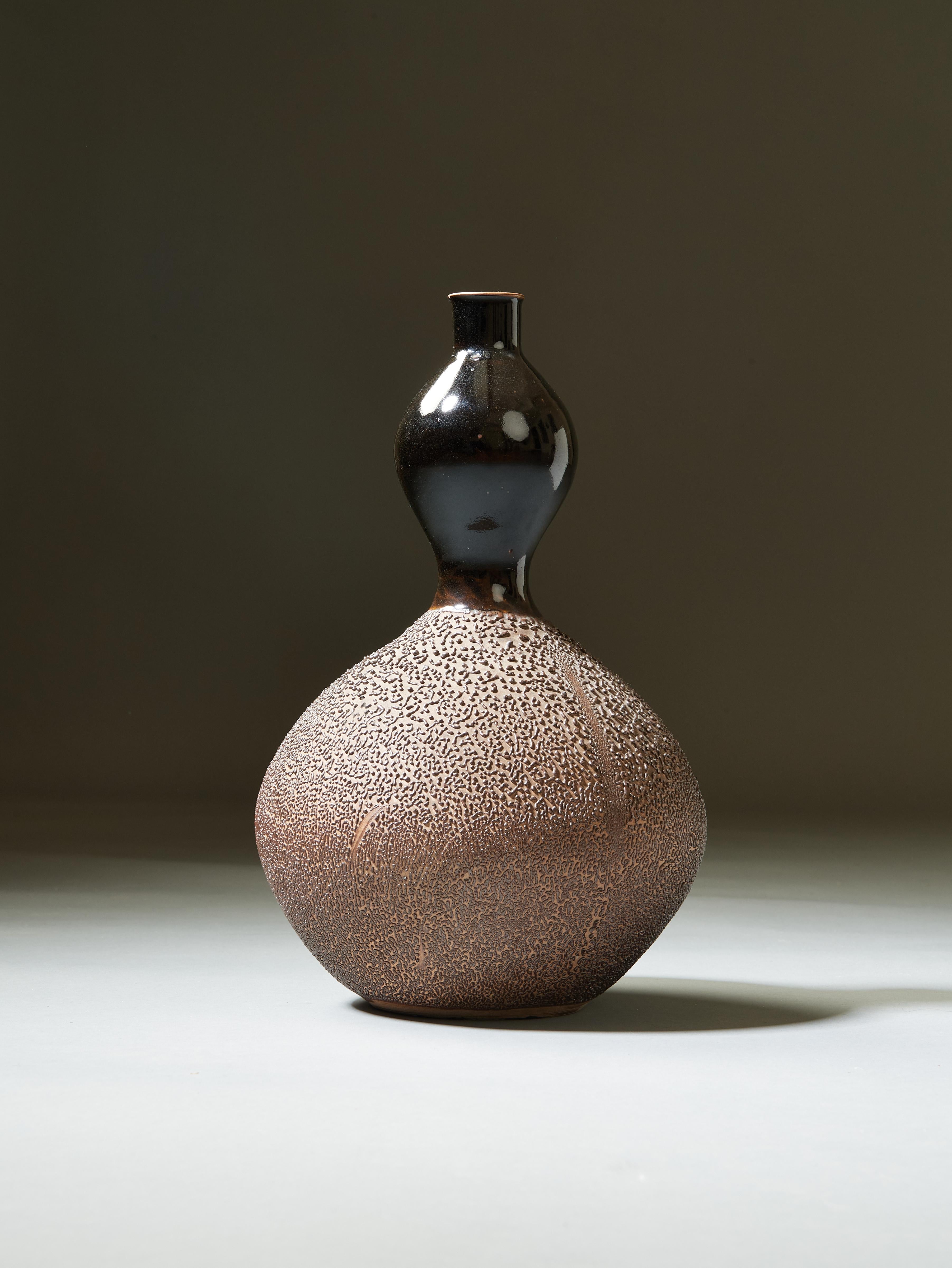 Japan, XXth Century

A sinuous Japanese double-gourd vase, in glazed black and textured earth-toned stoneware. A compressed globular lower bulb, its matte, beautifully tactile surface mottled like melon rind, flows into a smaller upper bulb, with a