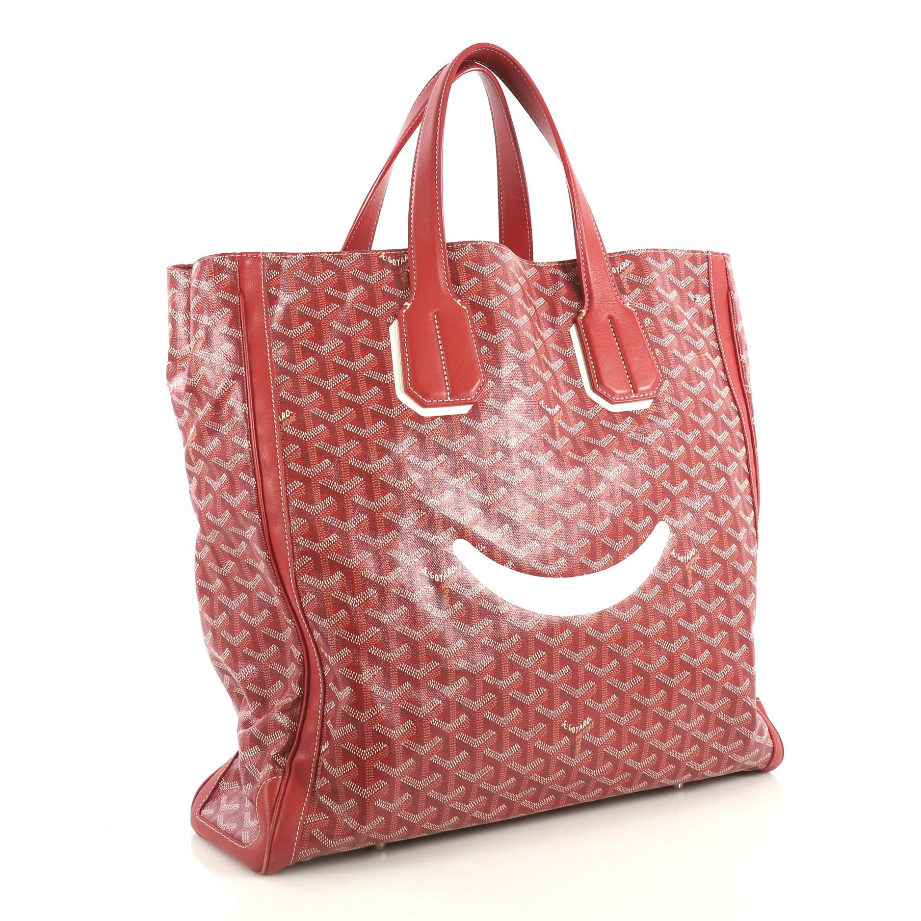 This Goyard Voltaire Convertible Tote Painted Coated Canvas, crafted from red coated canvas, features dual flat leather handles, protective base studs, smiley face accent at front, and silver-tone hardware. Its open top with middle hook closure