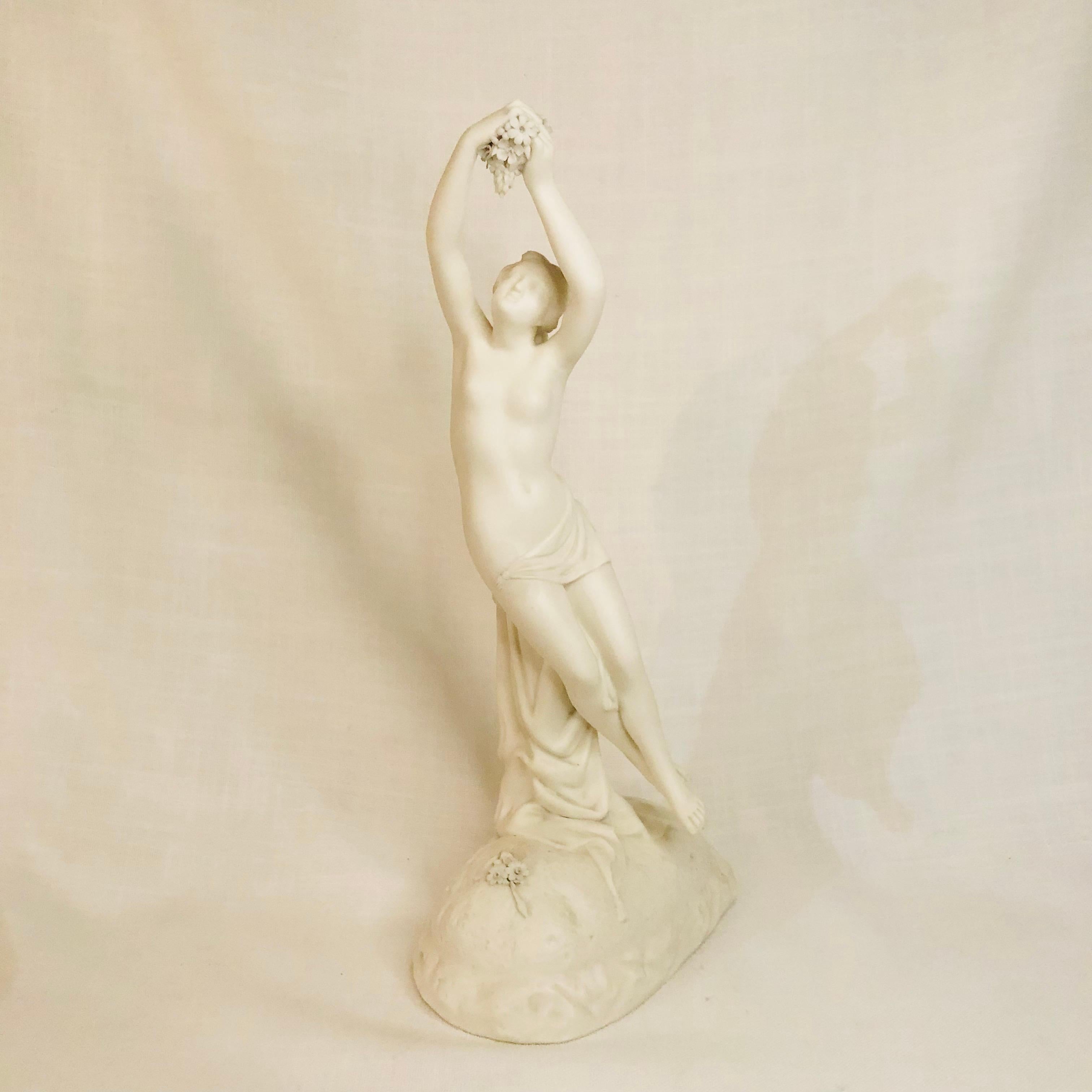 This is a very graceful and beautiful English parian figure of a semi-nude lady who is looking upward at the grapes she is holding with her outstretched arms above her head. She has draped flowing fabrics around her lower body. This figure is made