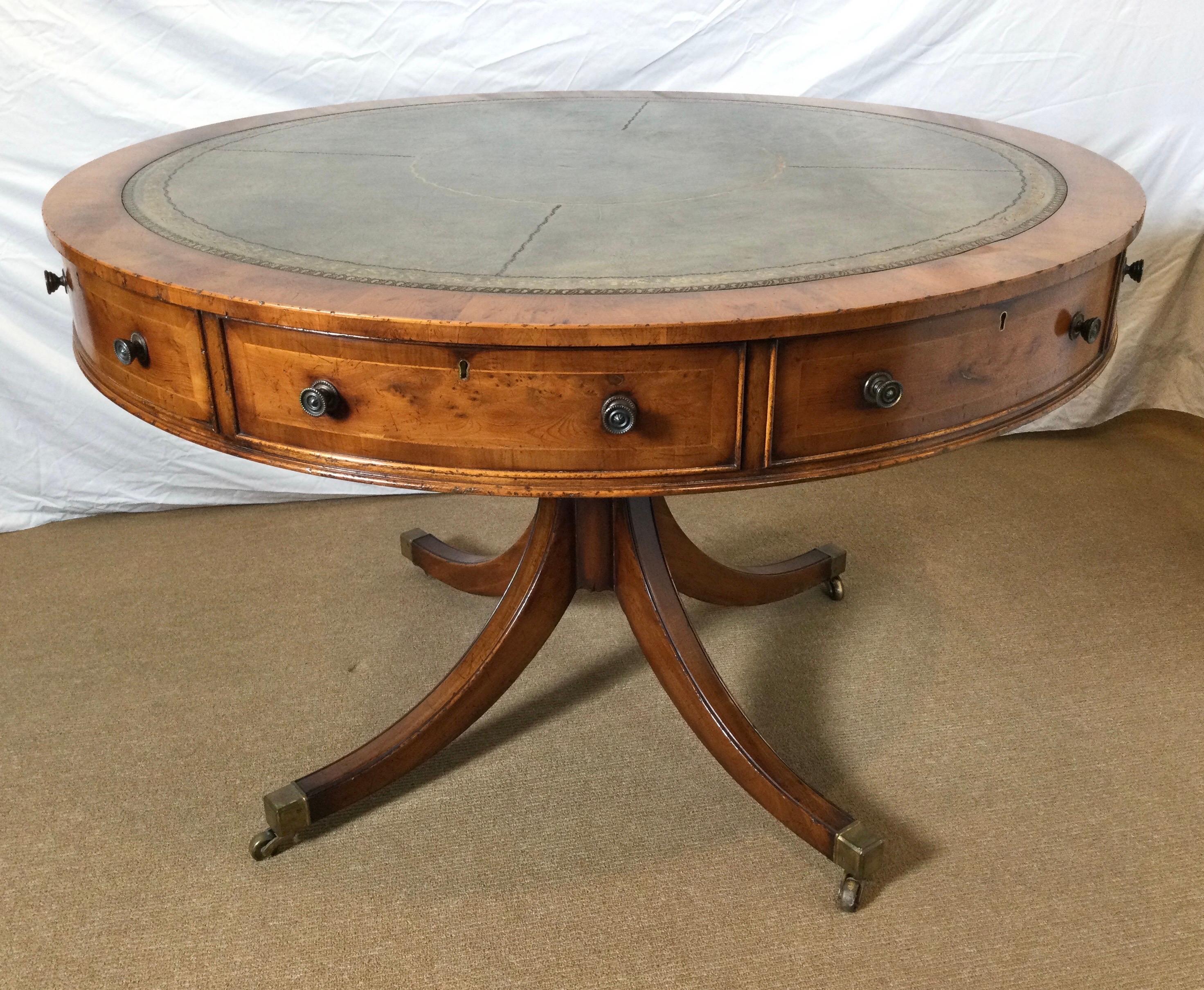 A classic round drum tale with medium olive tooled leather top. The thick apron with four working drawers and four fixed drawer fronts on a pedestal base with four splayed legs resting on brass castors. The wood is a smoky medium color Yew wood with