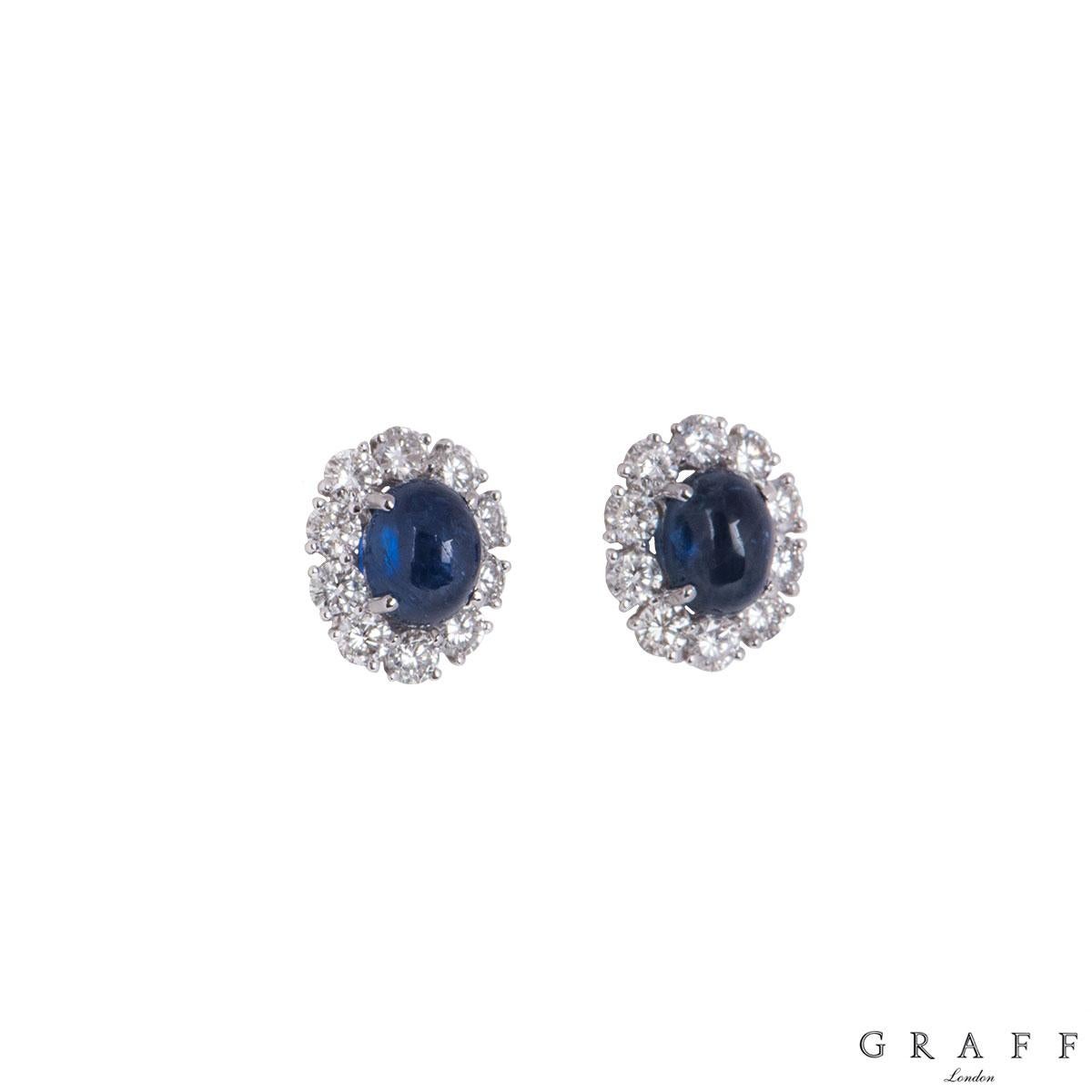 An 18k white gold pair of diamond and sapphire earrings by Graff. The earrings comprise of an oval shaped blue sapphire in the centre within a four claw setting with a halo of 10 round brilliant cut diamonds. The sapphires have an approximate total