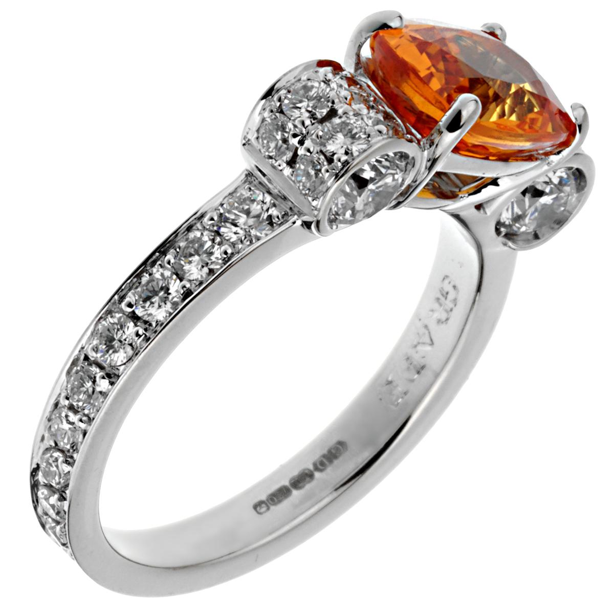 An incredible Graff ring showcasing a 2.45ct cushion cut Orange Sapphire flanked by 1.53cts of the finest round brilliant cut diamonds in 18k white gold. The ring measures a size 6 1/2 and can be resized if needed. The Orange sapphire very bright
