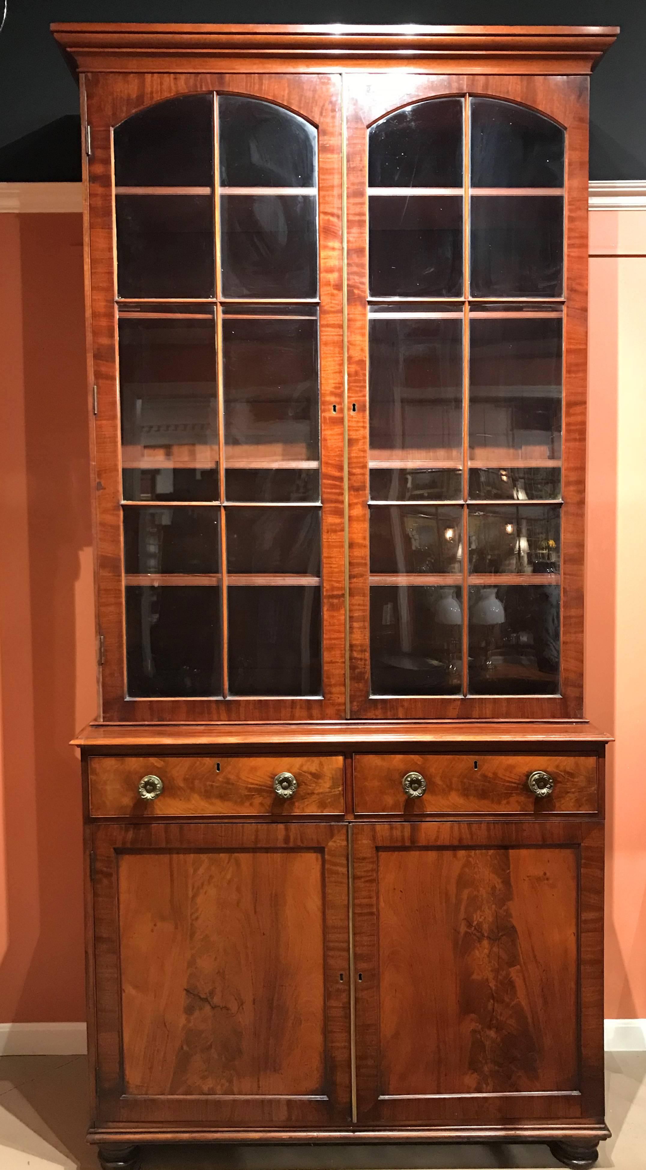 A beautiful large English two-piece mahogany step back bookcase, its upper case with a molded cornice over two glass doors, each with ten glazed glass panes, the top two panes arched on each, opening to four adjustable interior shelves, surmounting