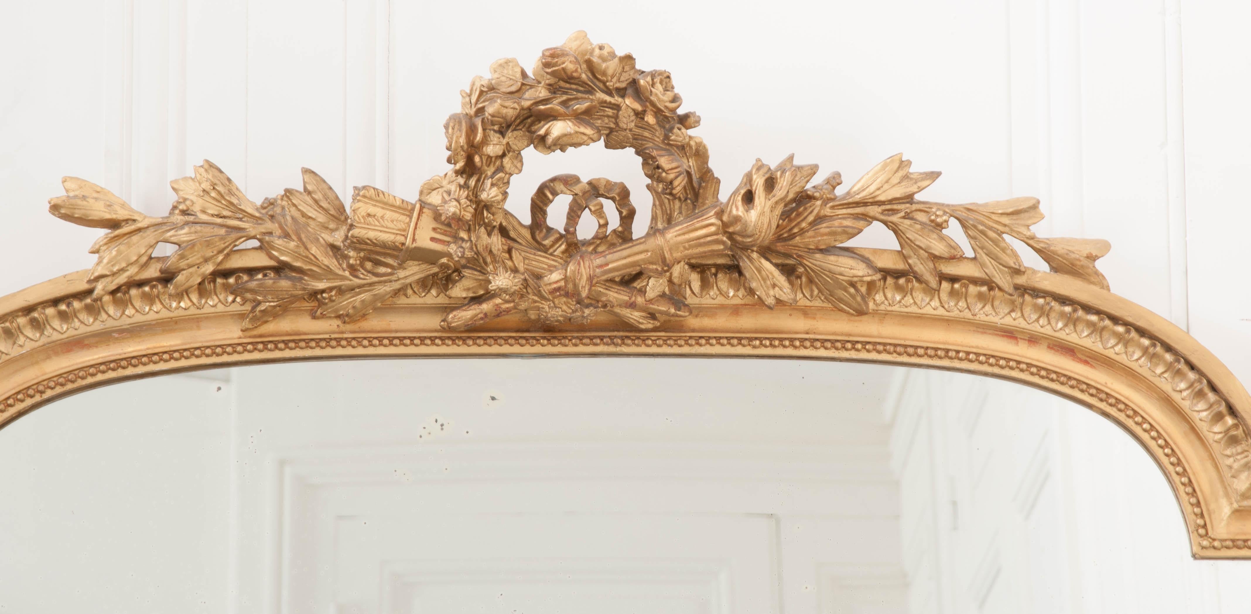 A spectacular gold gilt mirror from 19th century France. An impressive crest rests atop the mirror’s shaped top rail. This crest is composed of a wreath of roses set behind a crossed torch and quiver of arrows. A knotted bow can be seen in the