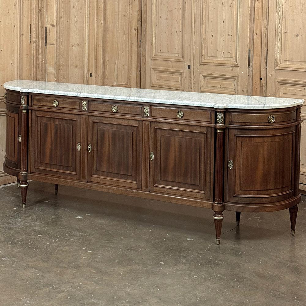 Il Grand French Louis XVI Mahogany Round-Sided Buffet with Carrara Marble è un magnifico esempio del revival classico avvenuto a metà del XX secolo in Francia.  Dotato di ben cinque cassetti e cinque ante, vanta lati curvi ad ampio raggio per
