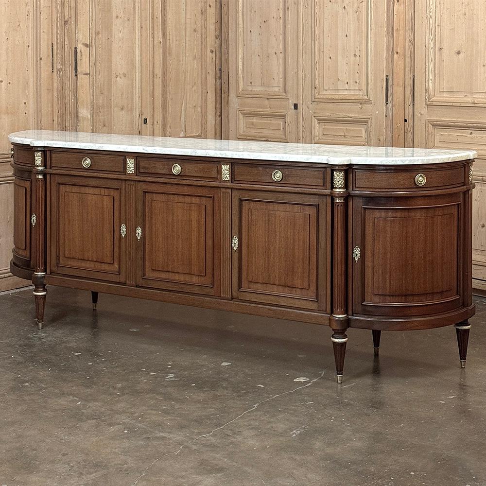 Il Grand French Louis XVI Mahogany Round-Sided Buffet with Carrara Marble è un magnifico esempio del revival classico avvenuto a metà del XX secolo in Francia.  Dotato di ben cinque cassetti e cinque ante, vanta lati curvi ad ampio raggio per