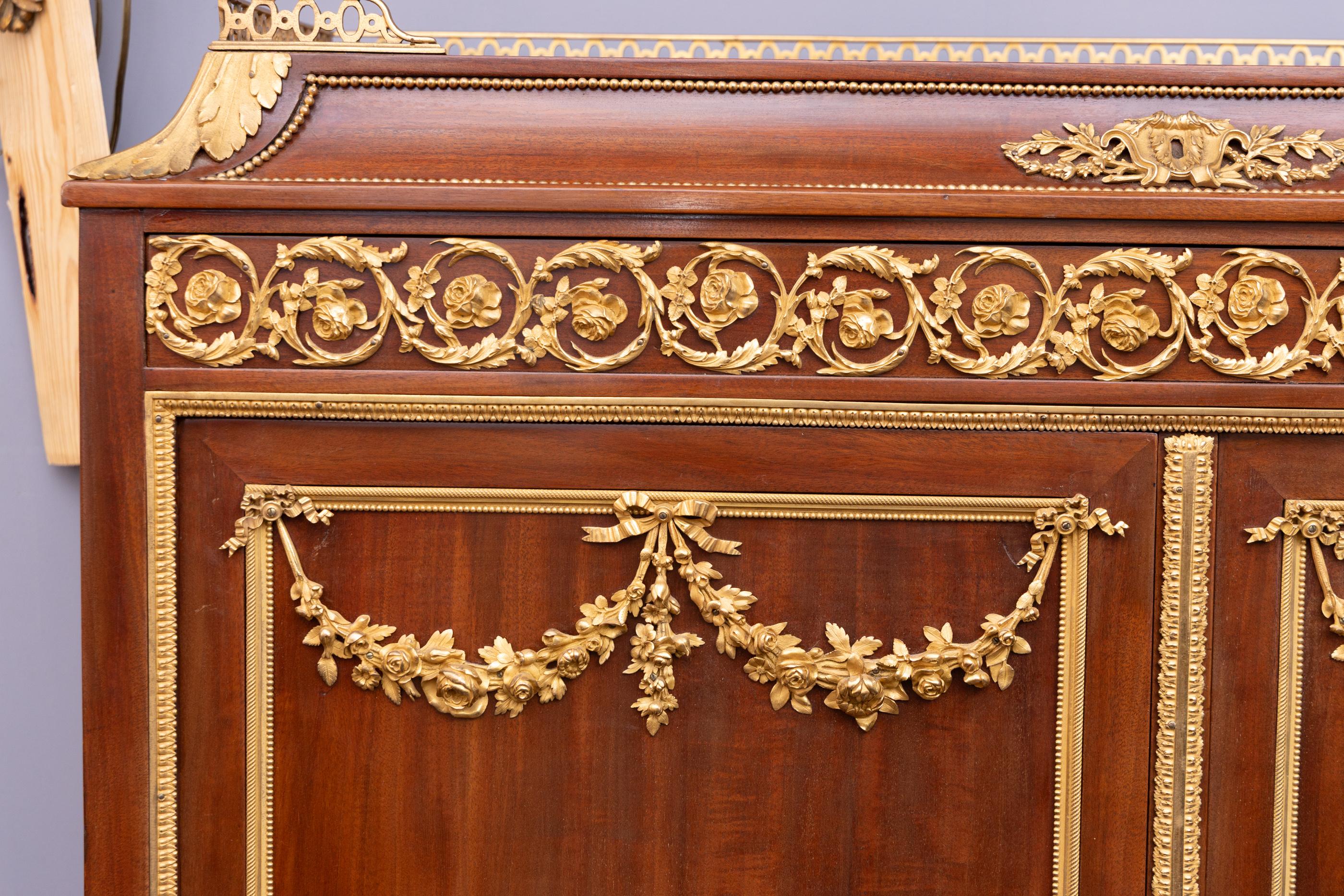 Questo mobile Luigi XVI a quattro ante in mogano è impreziosito da festoni e decorazioni in bronzo d'ore;  Sulle porte ci sono delle volute floreali con un motivo a fiocco al centro.  Tra le grandi porte superiori e quelle inferiori si trova un