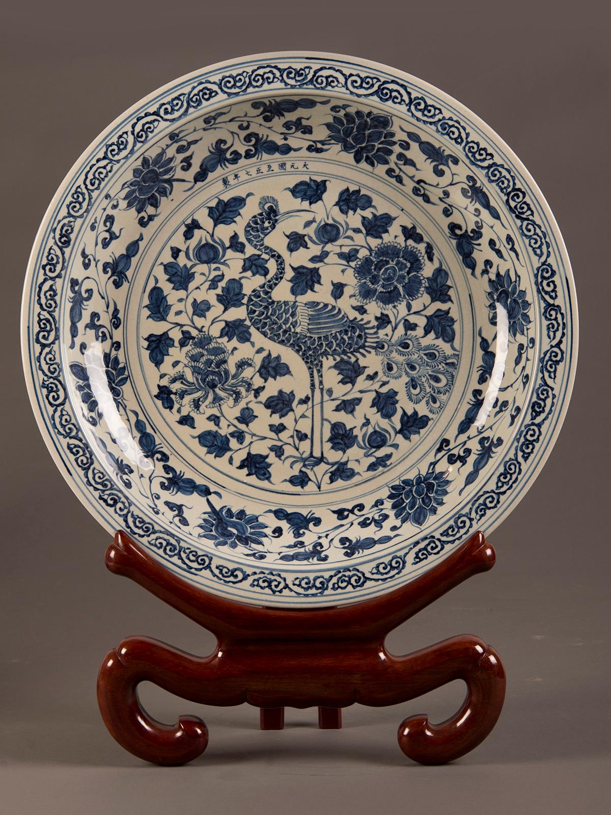 A grand scale hand painted Chinese blue and white glazed shallow bowl from China. The striking image of the crane with its distinctive stance amidst a floral fantasia is the focal point of this large piece displayed on a substantial rosewood colored