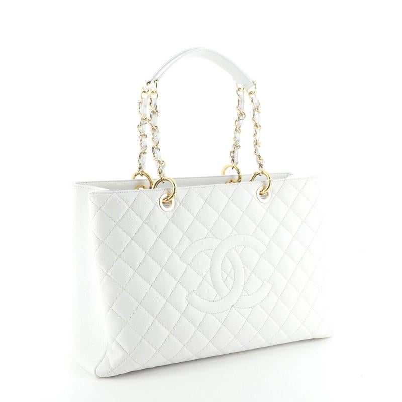 This Chanel Grand Shopping Tote Quilted Caviar XL, crafted in white quilted caviar leather, features woven-in leather chain straps with leather pads, stitched CC logo, exterior back pocket, and gold-tone hardware. It opens to a gray satin interior