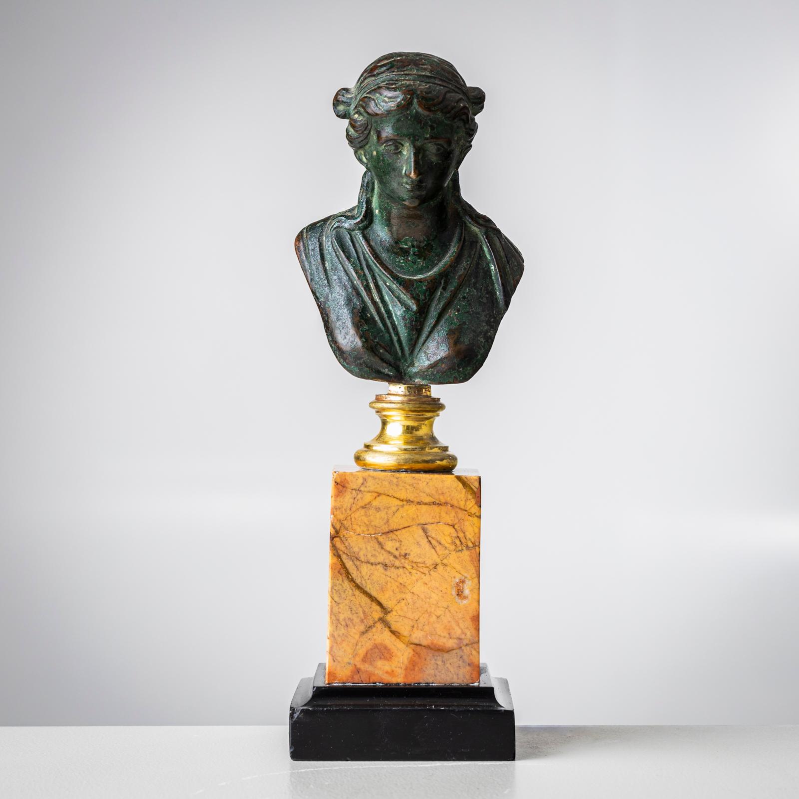 Busto femenino de bronce de mujer con pie dorado, que descansa sobre un plinto de mármol marrón con base de madera negra. Pátina natural. Zócalo 7 x 7 cm