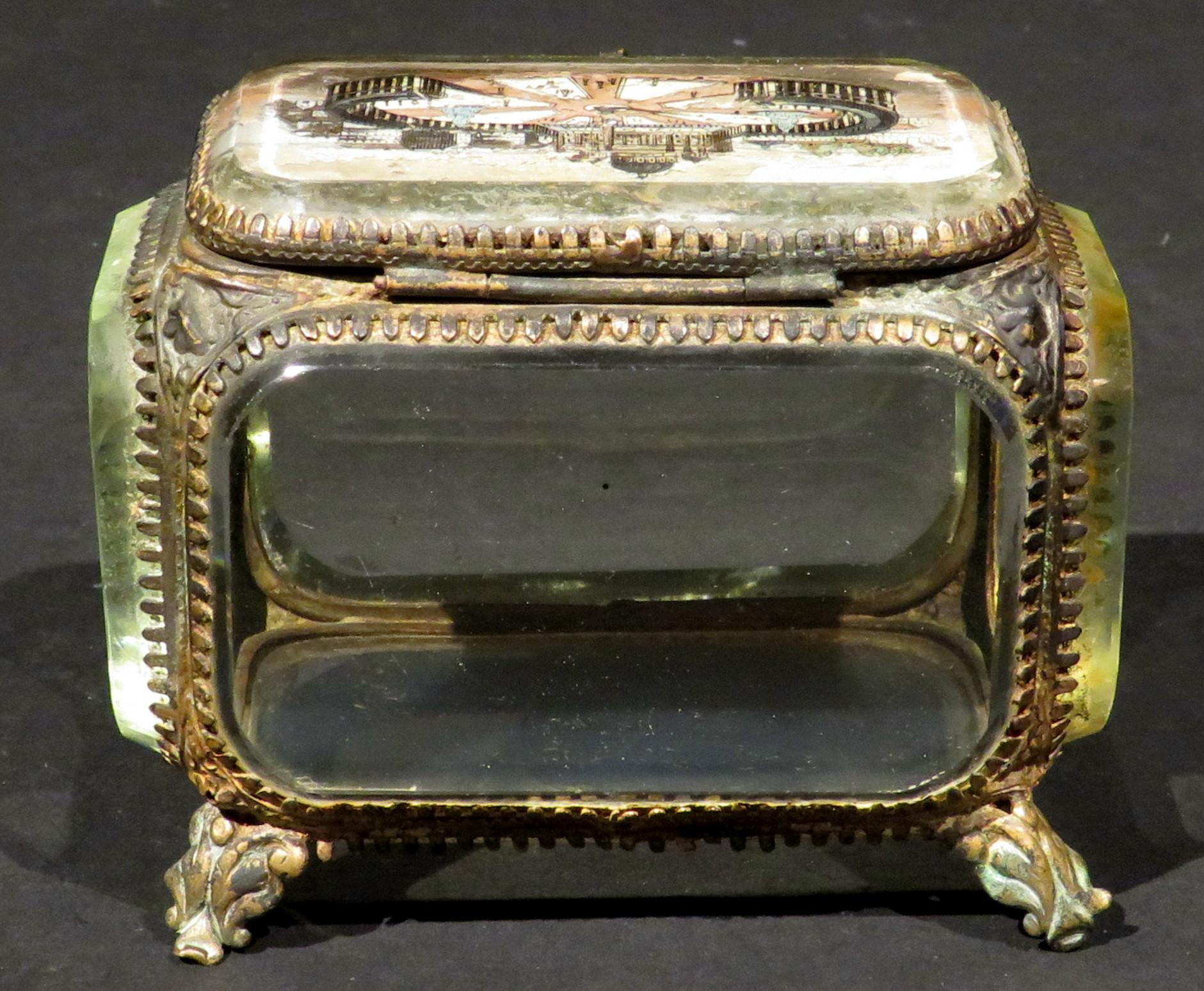 A diminutive Grand Tour style glass trinket box constructed of bevelled glass panels set within an ormolu case on all sides, raised overall upon foliate shaped feet. The hinged top featuring a reverse painted view of Saint Peter's Square in Rome. No