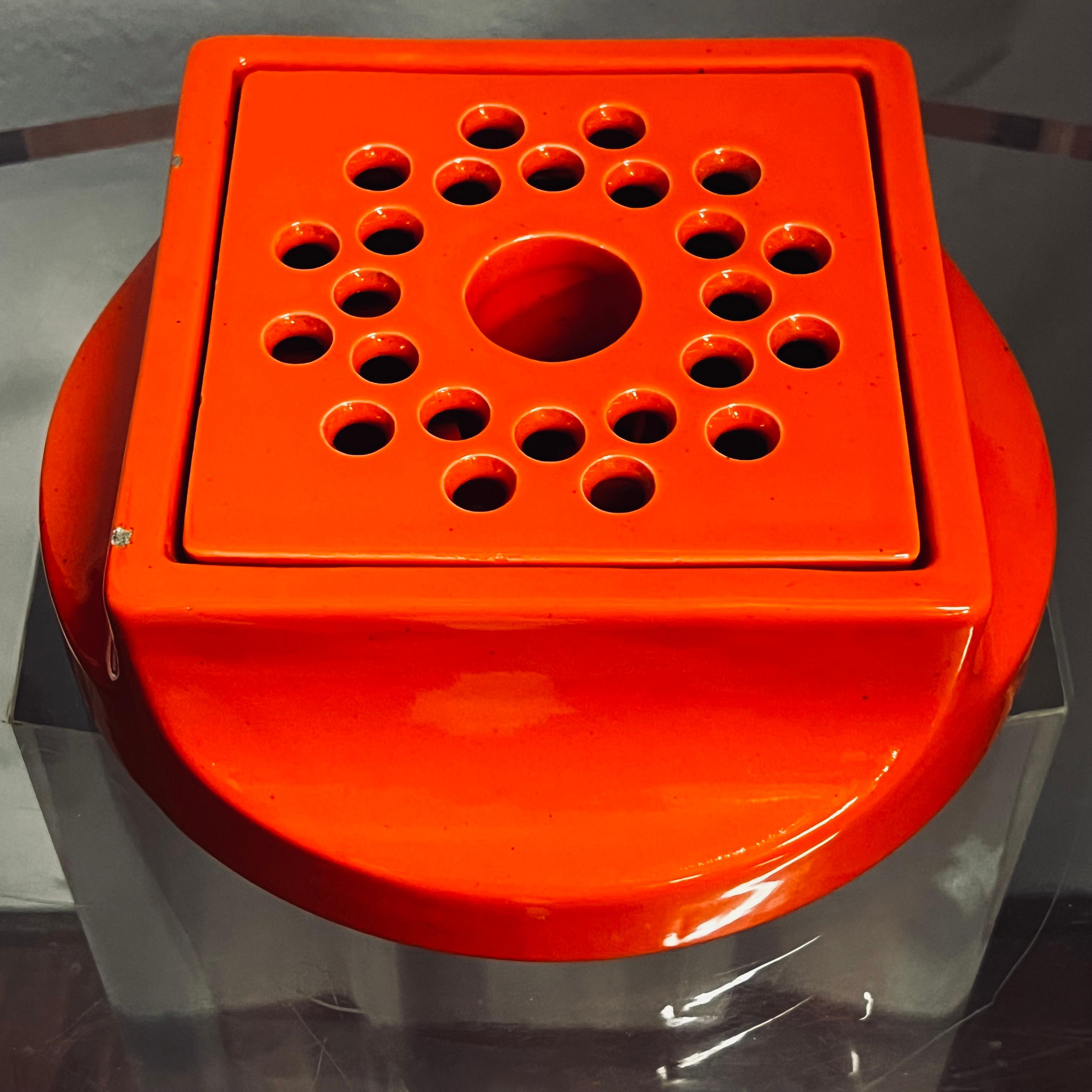 Grande posacenere in ceramica arancione, disegnato dallo Studio OPI per Gabbianelli, composto da un corpo contenitore tondo che sviluppandosi verso l'alto, diventa quadrato accoglie una griglia forata, anch'essa in ceramica che sul retro riporta le