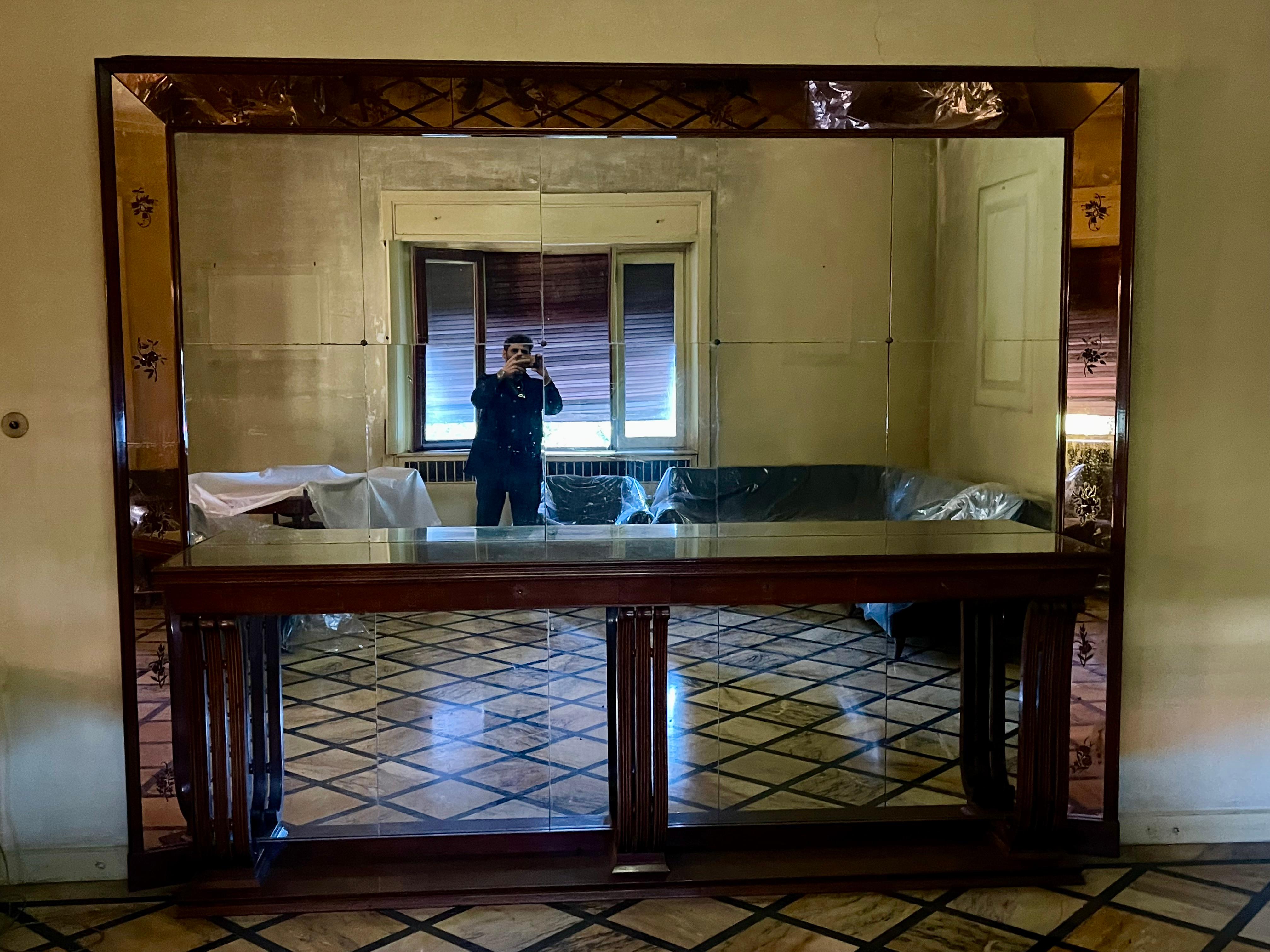 We are looking at an imposing hall or living room mirror with wooden drawer console.
The dimensions are significant, measuring 3 meters wide by 2.45 meters high.
It consists of a large wooden plank with a carved frame, on which are housed a series