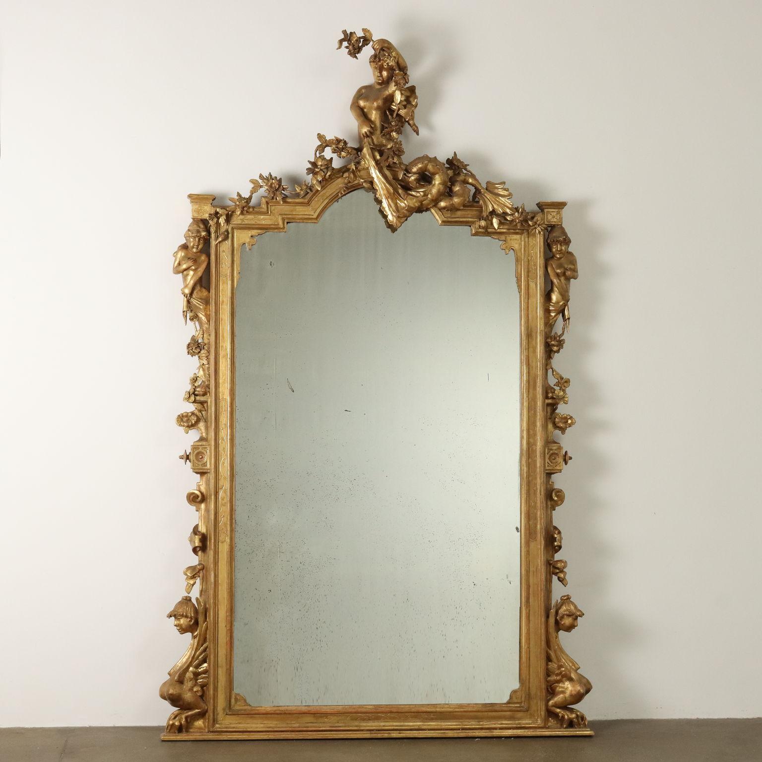Monumentale specchiera d'appoggio, 1870ca. Cornice incisa a bulino contenente specchio a cui si appoggiano i ricchi intagli, nella base due sfingi alate si contrappongono mentre al vertice dei montanti altre due cariatidi reggono un festone di fiori