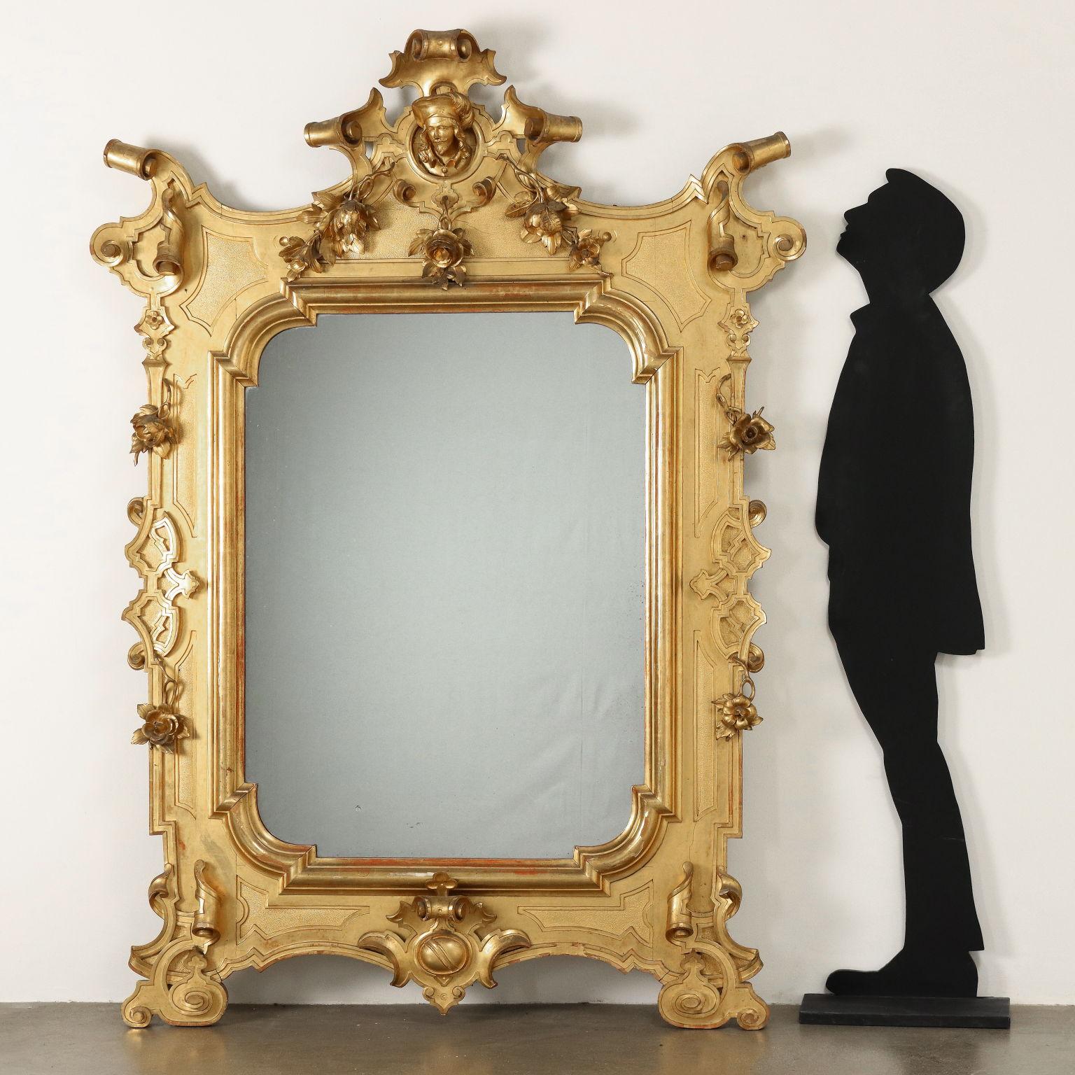 Grand miroir d'époque ombertine. Sculptée de grands rinceaux, de boucles et de lambrequins superposés, dans la cimaise un médaillon avec une tête de chevalier et des branches de roses sculptées, miroir au mercure, toute la surface est dorée à la