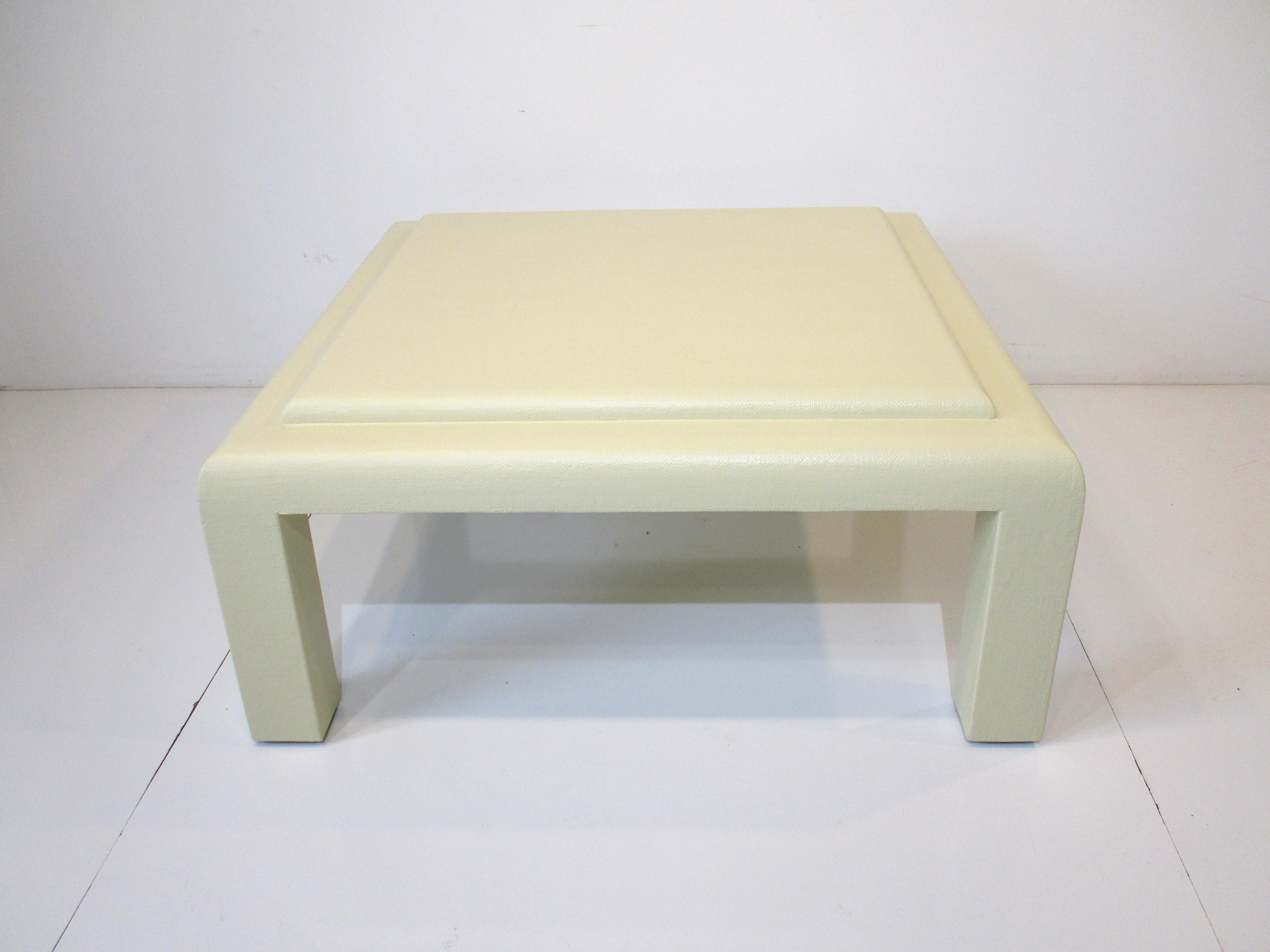 A mid toned cream finished grass cloth covered coffee table with tiered upper center top . Rounded edges to the top with nice design contracting sharper edged legs in the manner of Karl Springer.