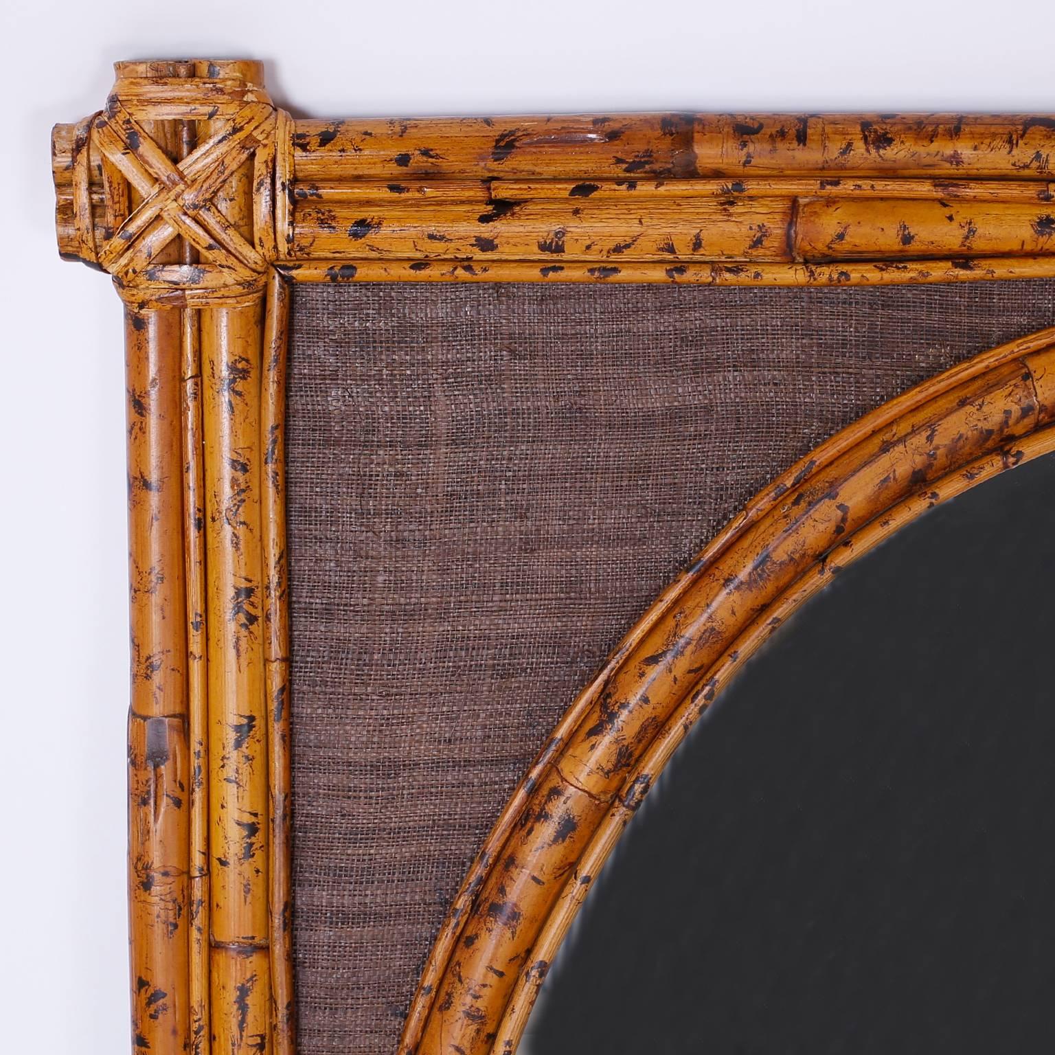 Oval mirror in a casually elegant rectangular frame crafted with bamboo, burnt for effect, and wrapped with reed at the corners and having a grasscloth background.