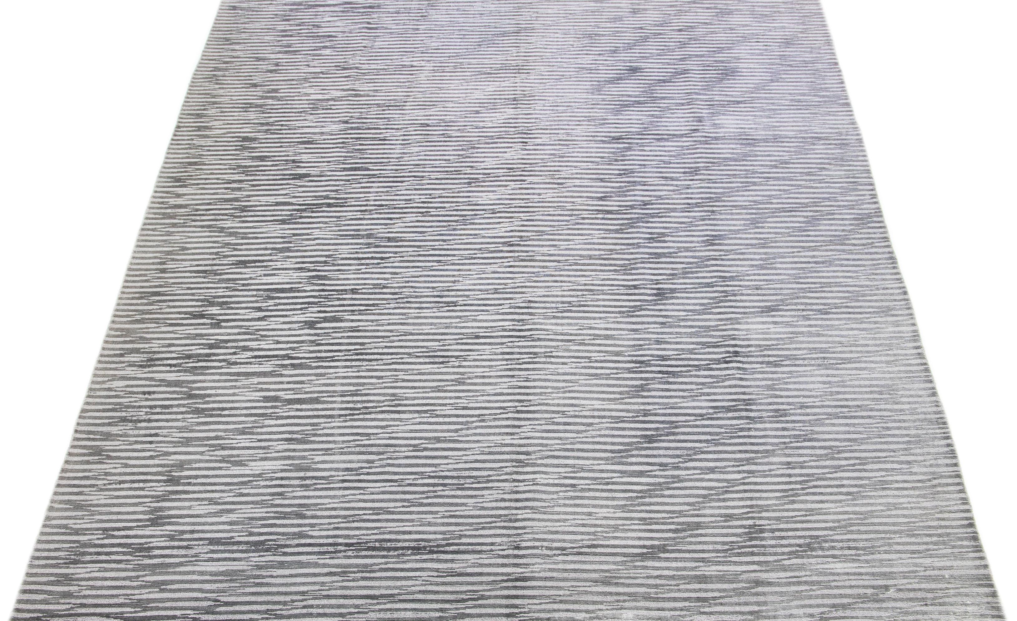 Dieser exquisite zeitgenössische Teppich, der in sorgfältiger Handarbeit aus einer Mischung aus Seide und Wolle gefertigt wurde, zeigt ein atemberaubendes graues Grundmuster, das durch ein verführerisches Streifendesign in Silbernuancen perfekt