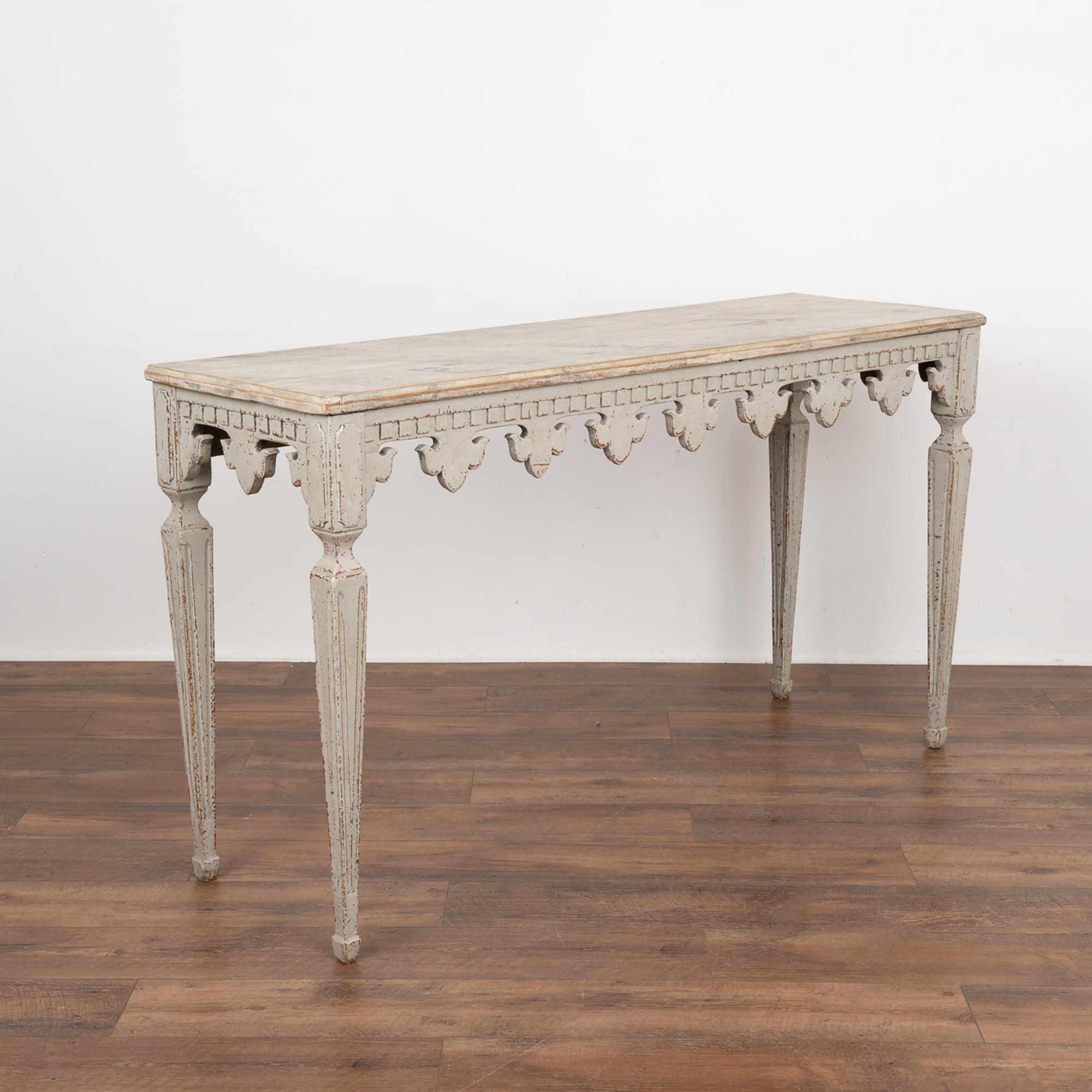 Elegant console table with carved fleur de lis skirt topped by dentil molding, raised on graceful tapered fluted legs.
Later applied professional painted finish in layers of gray with contrasting faux marble top. The finish has been lightly
