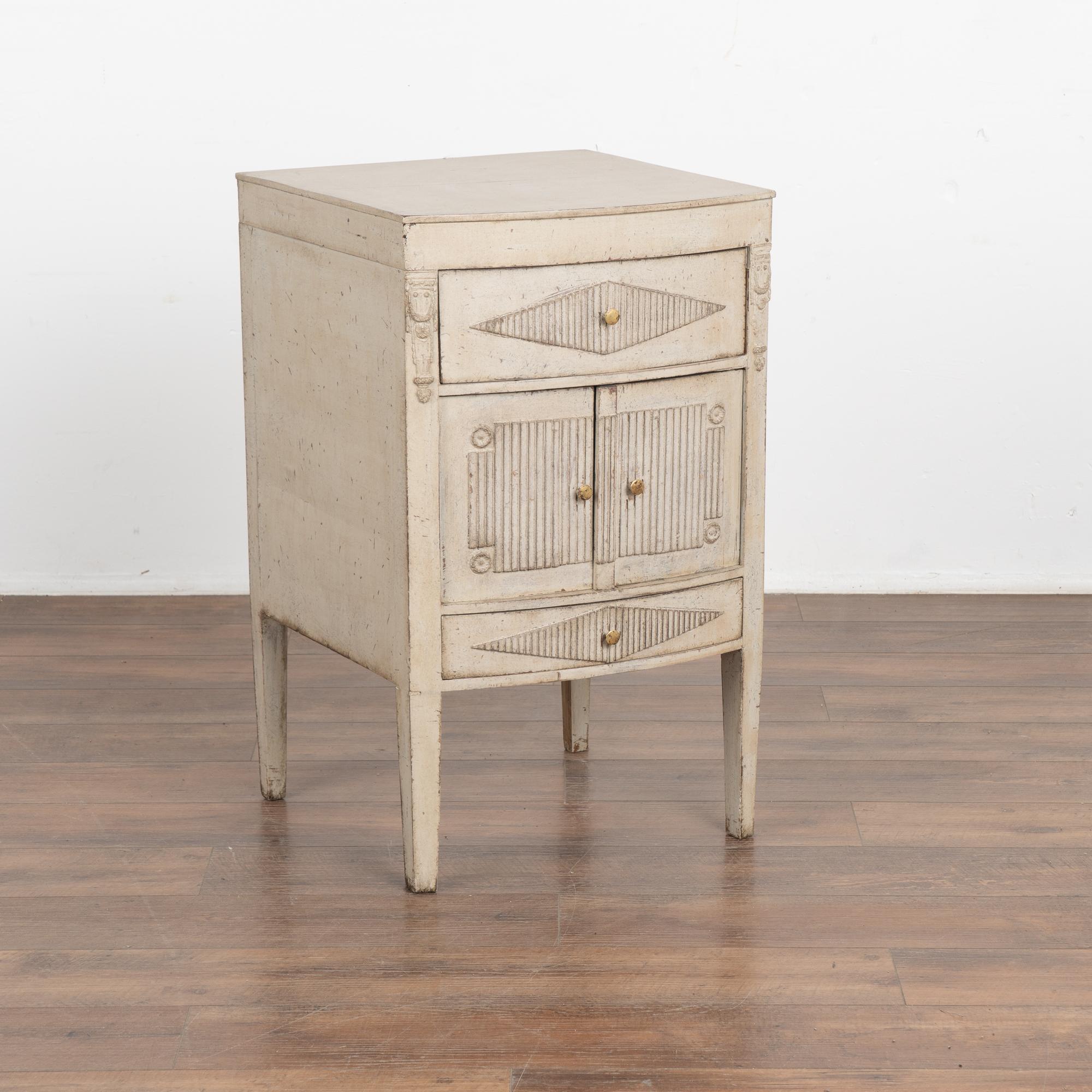 Small Swedish country pine cabinet standing on tapered legs; may serve also as a nightstand or side table.
Small drawers with traditional fluted diamond accent sits above and below the two small cabinet doors, each with a simple brass pull knob.
The