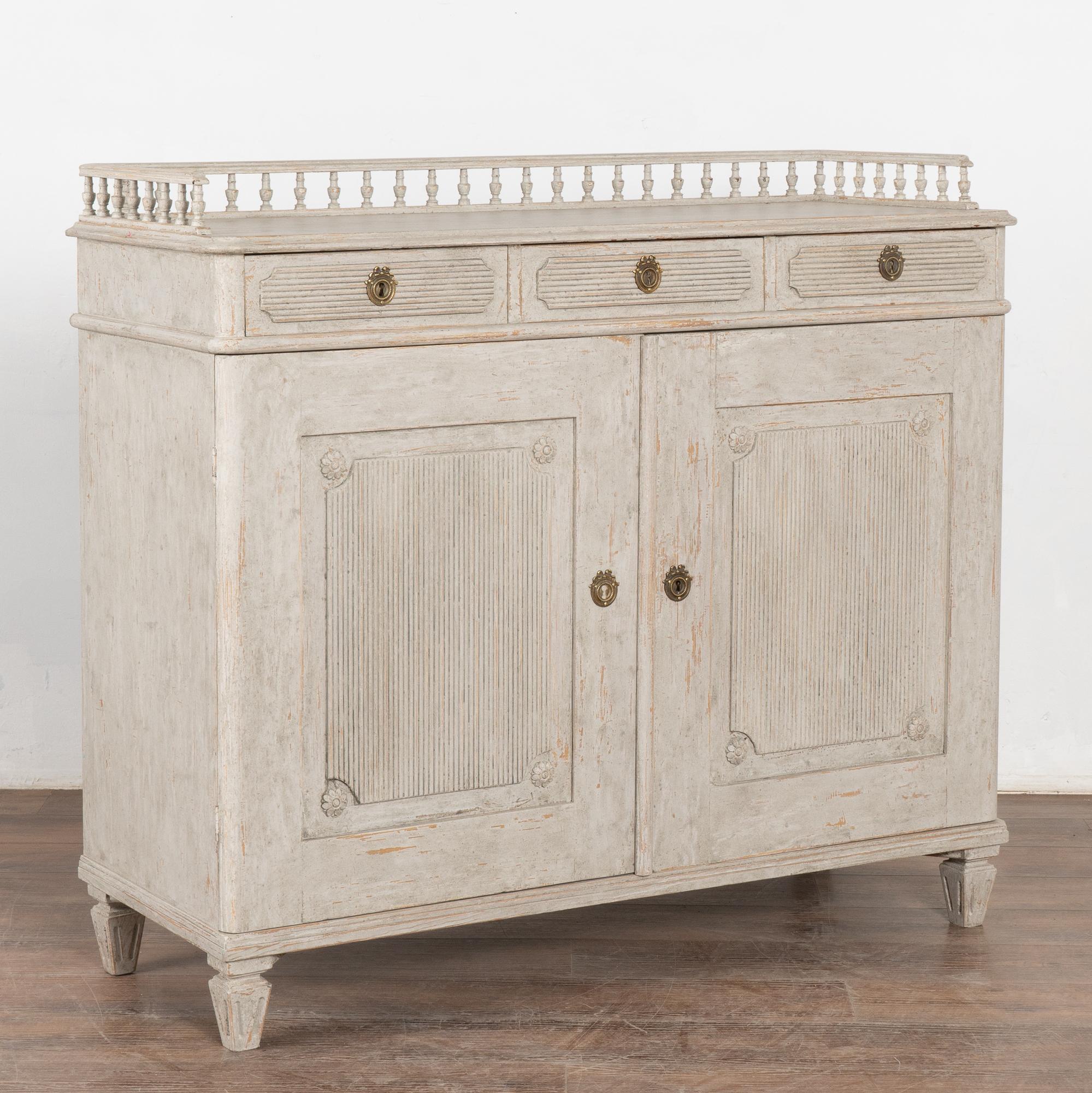 This Swedish Gustavian style sideboard has a (newer) soft gray painted finish (with white undertones) which complements the traditional fluted carving along the panel doors, drawers, and fluted tapered feet.
Two of the three drawers still have a