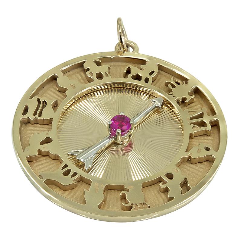 Large round charm, with full calendar of cut-out astrological signs around the inner border.  The center has a rayed line pattern, with a moveable arrow, set with a faceted ruby.  Made by DANKNER.  14K yellow gold.   1 1/2