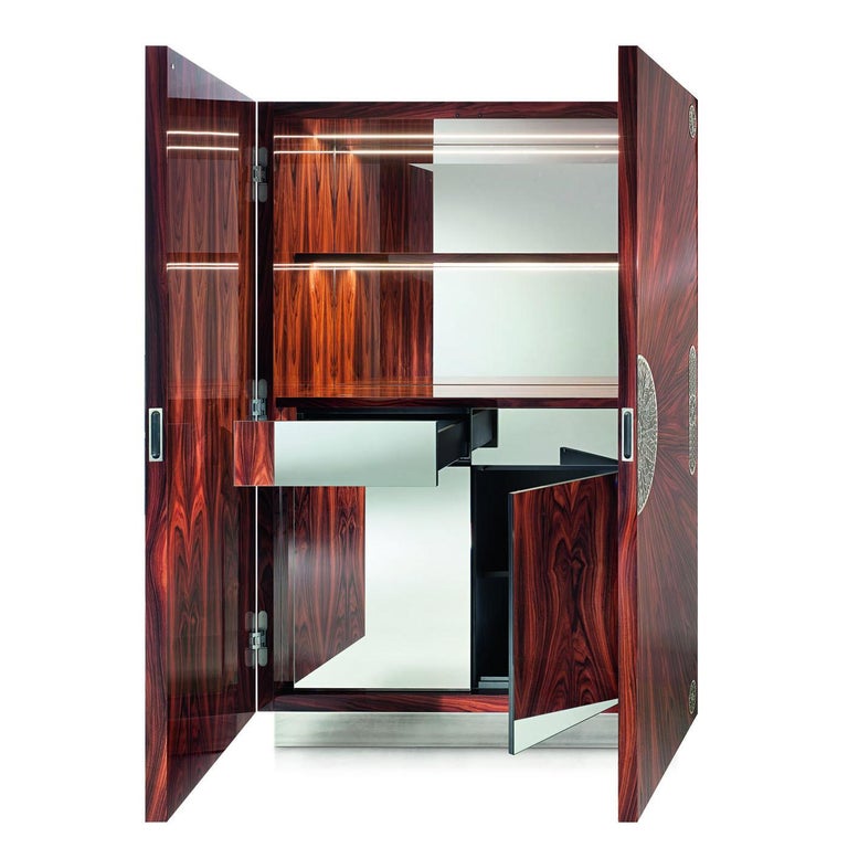Inspired by the style of the 20s with its geometric silhouettes and precious decoration, this cabinet bar mixes functionality and design to bring luxurious elegance to a living room. Its structure is in glossy-finished rosewood whose natural veins