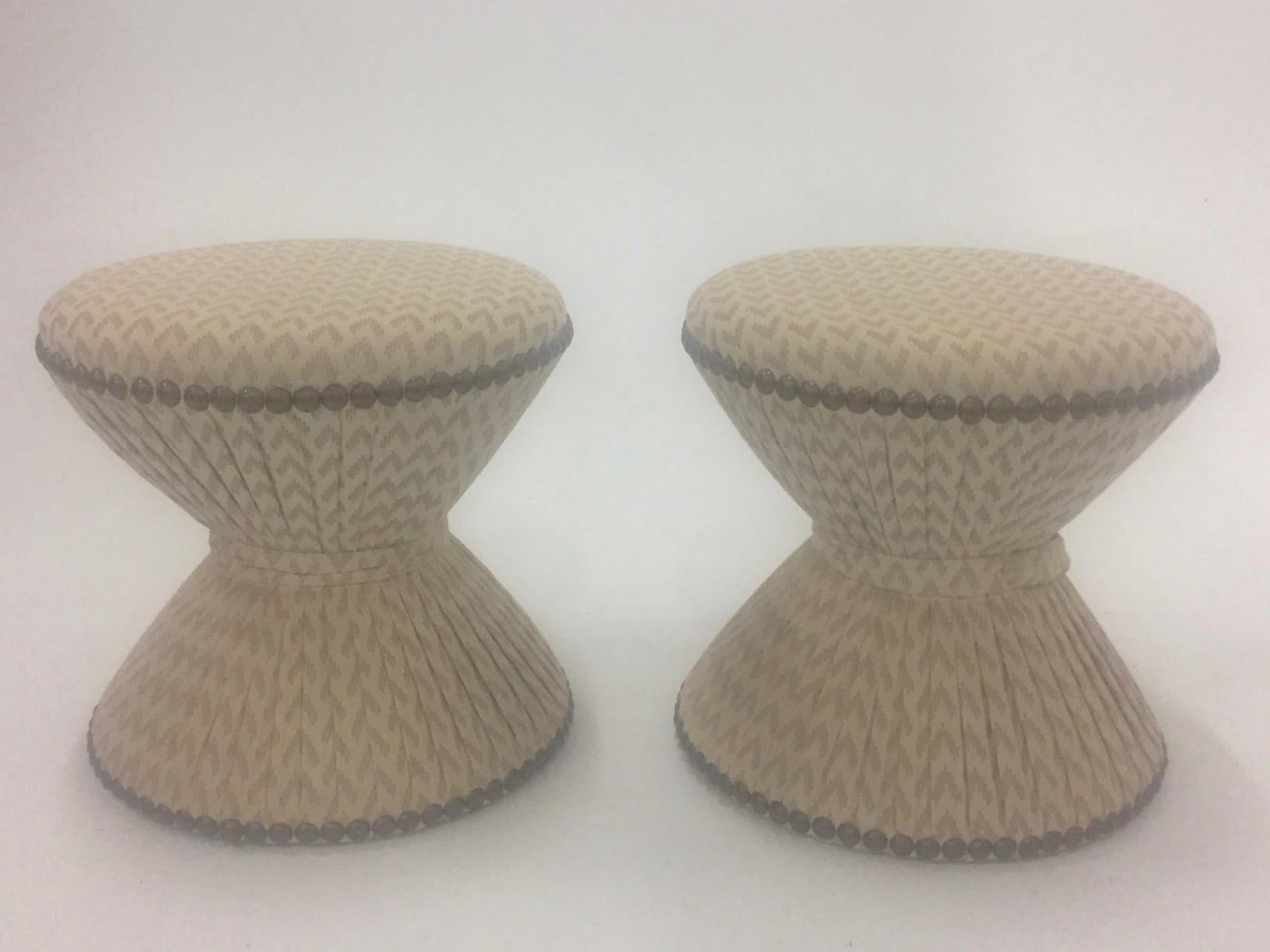 A pretty pair of fully upholstered hourglass shaped ottomans in a neutral cream fabric finished with nailheads.