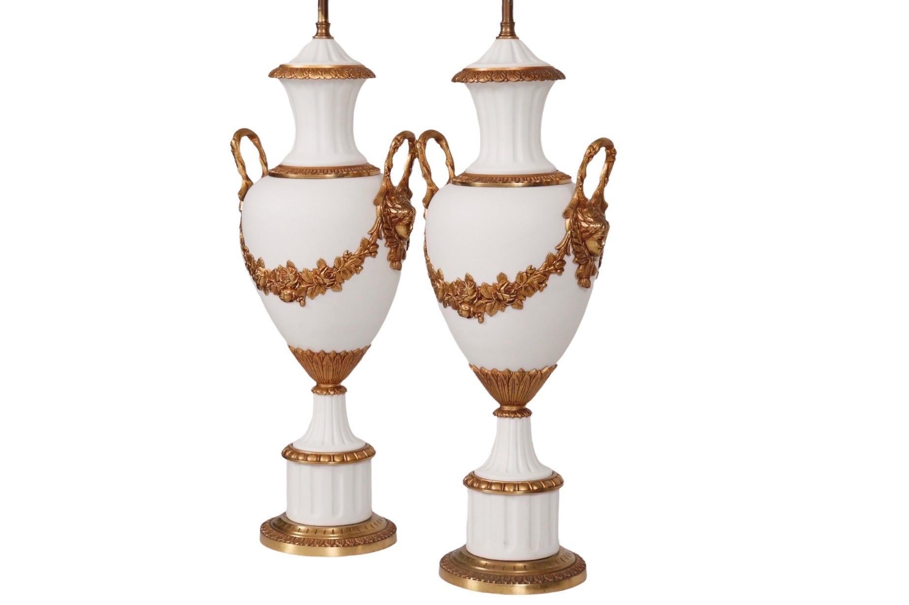 A pair of white bisque porcelain table lamps in the form of a Grecian urn, with fluting on the lid, neck and pedestal. Chased brass lotus leaves decorate the lid, base of the neck, round the lower body of the lamp and the pedestal base. Branch