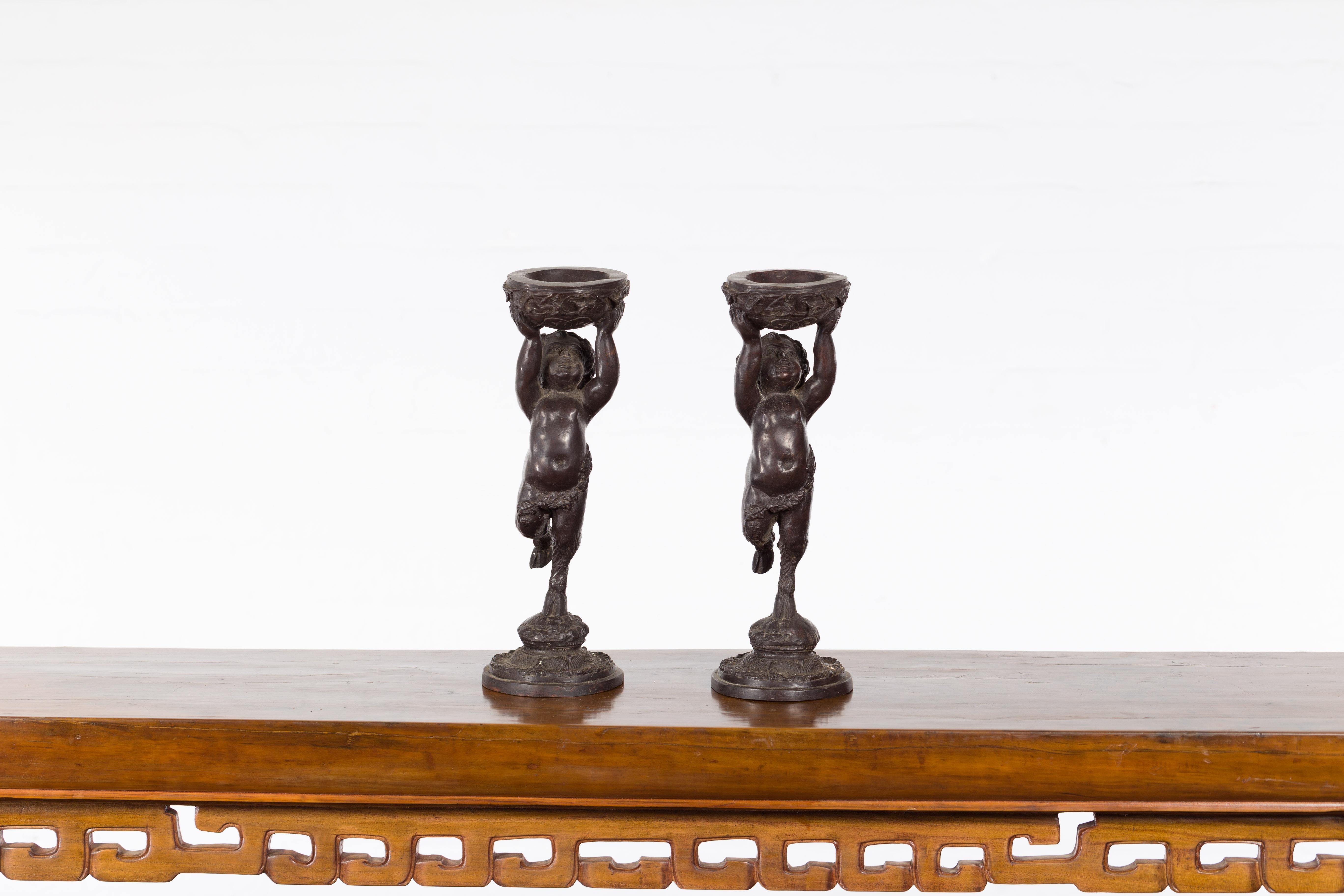 A pair of Greco-Roman style vintage bronze young satyr candle holders from the mid 20th century, holding circular vessels. Created with the traditional technique of the lost-wax (à la cire perdue) which allows for great precision and finesse in the